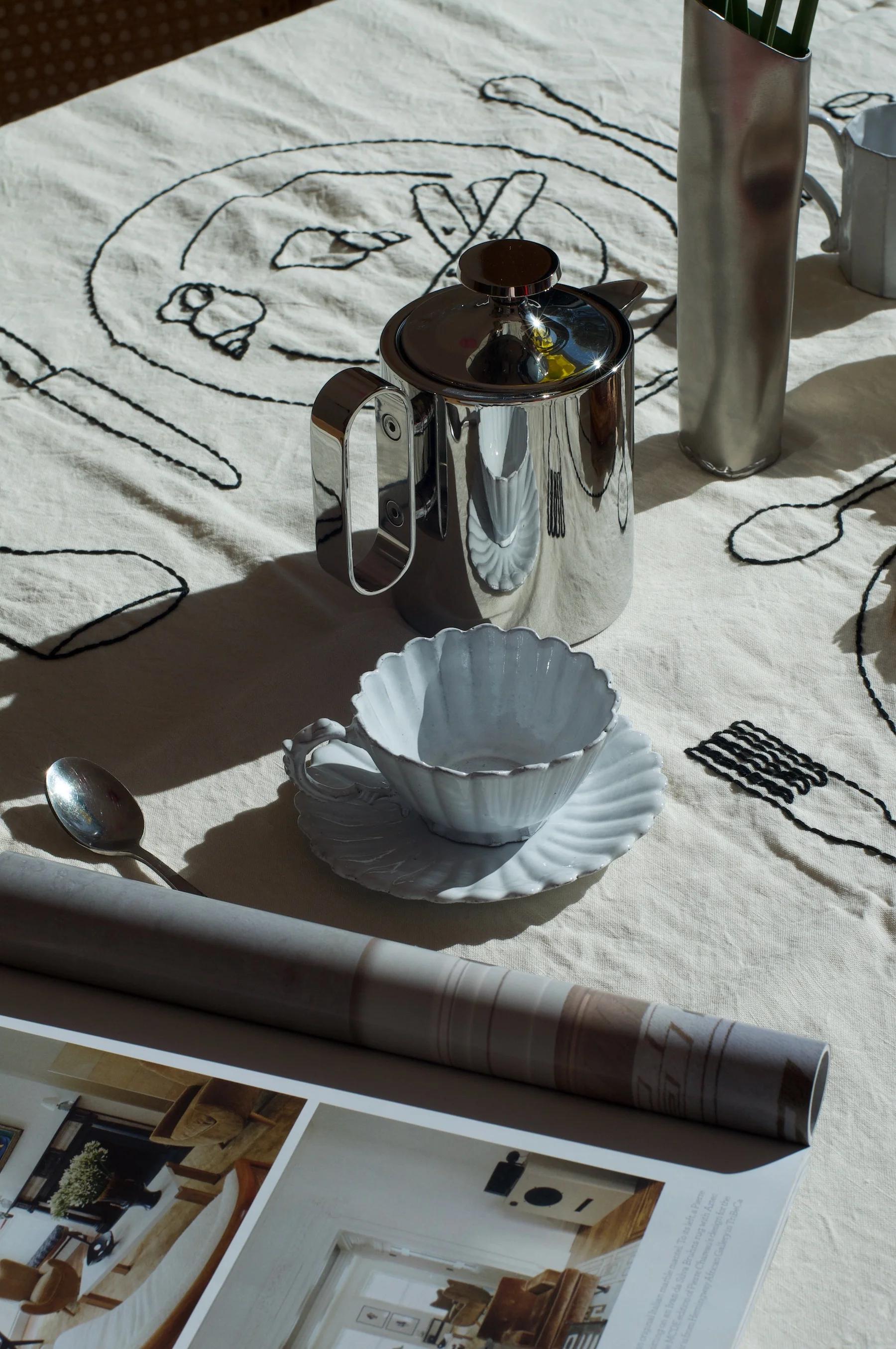 Table with embroidered cloth, metal teapot, white teacup and saucer, rolled magazine, open book. Metal vase with greenery.