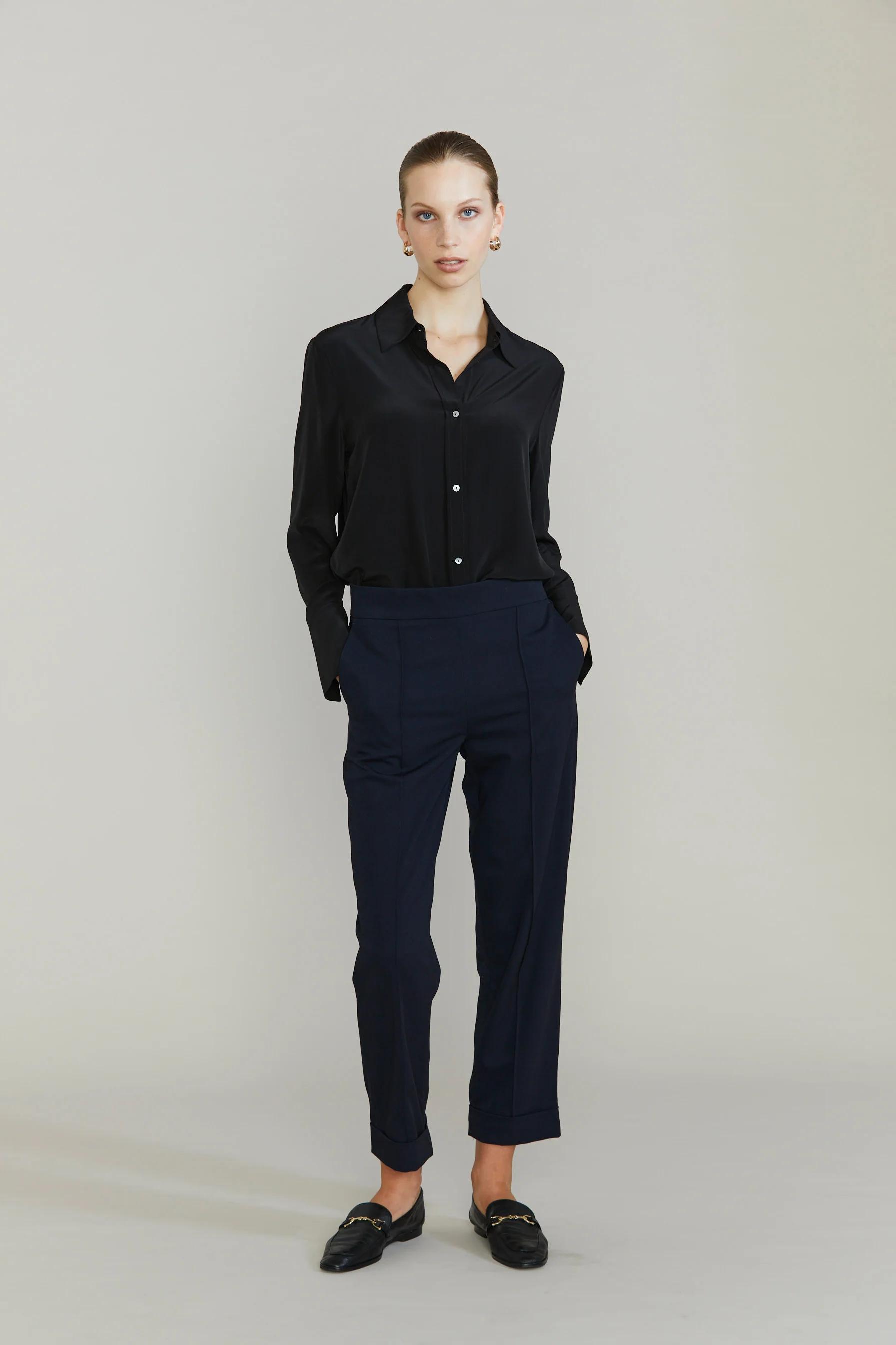 Person in black button-up shirt, navy pants, and black loafers. Hands in pockets. Plain background.