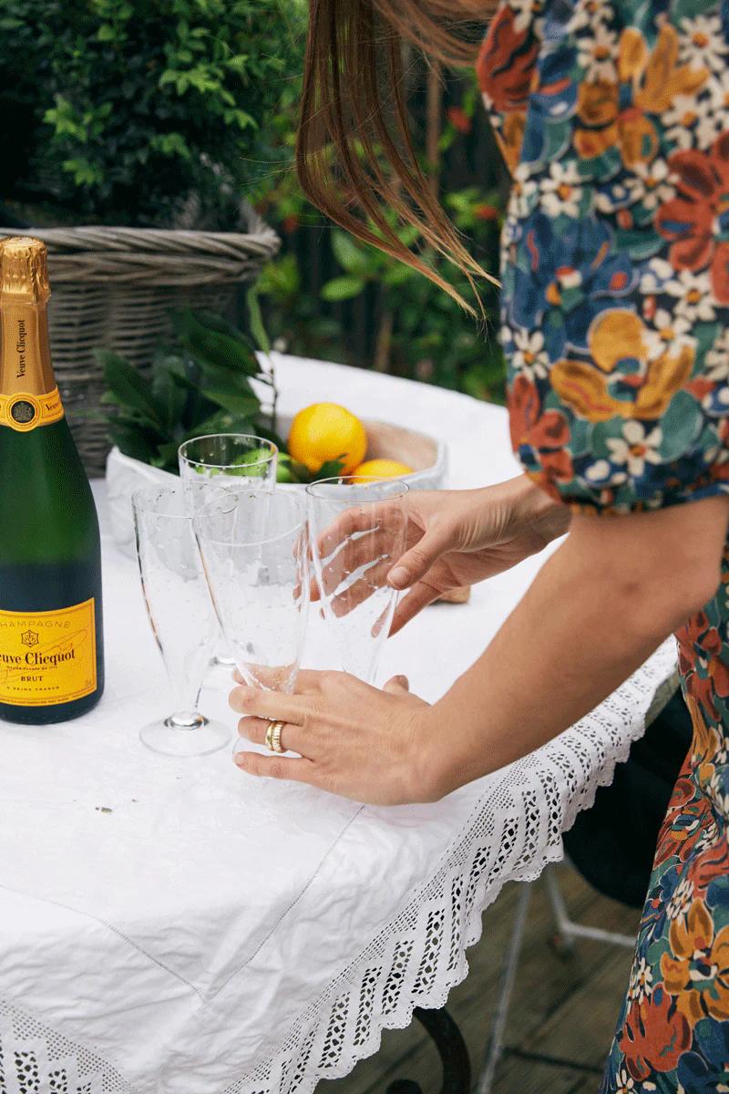 Person in floral dress sets champagne flutes on lace-covered table. Veuve Clicquot bottle, greenery, and lemons suggest outdoor celebration.