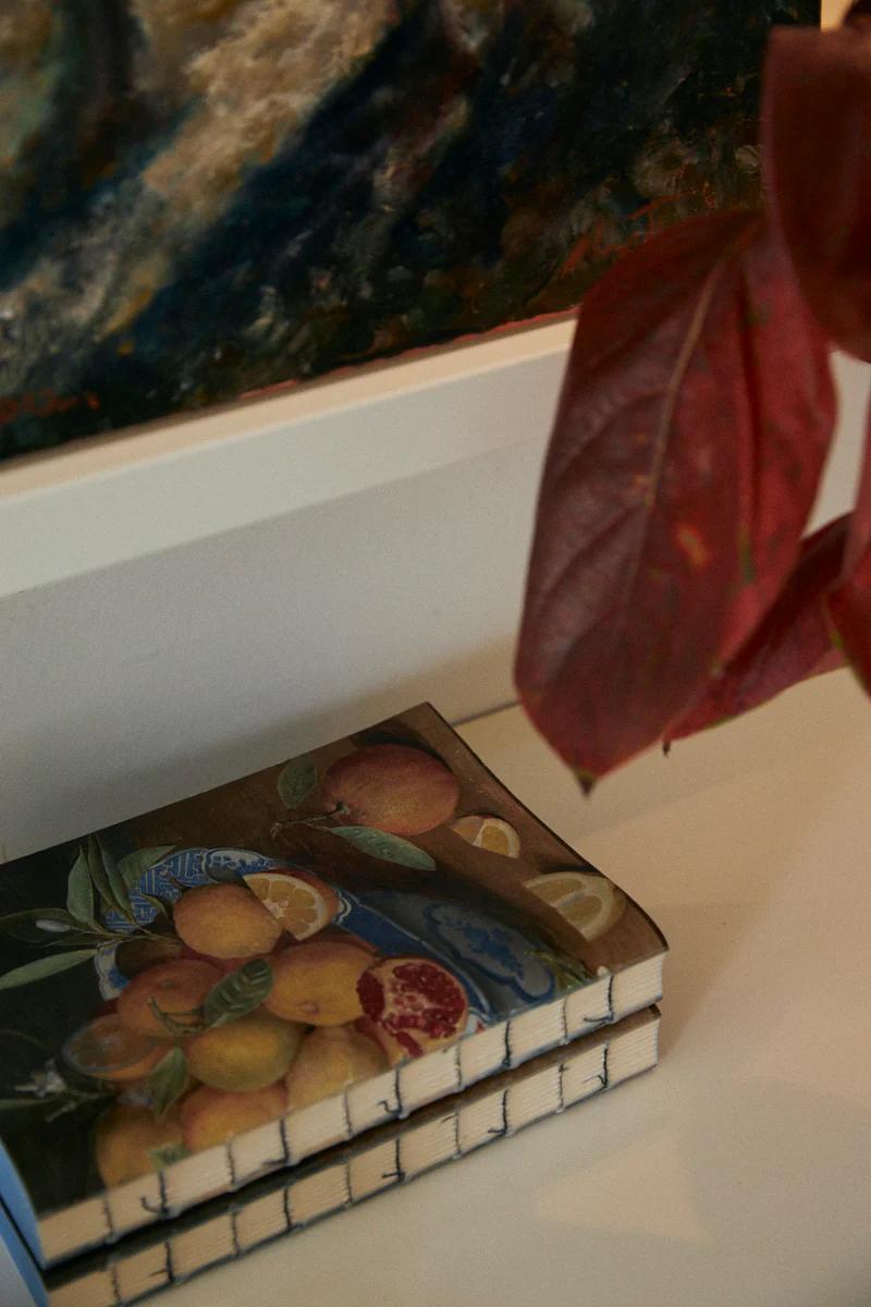 Fruit bowl painting on white shelf. Red leaf partly covers artwork. Textured canvas above shows craftsmanship.