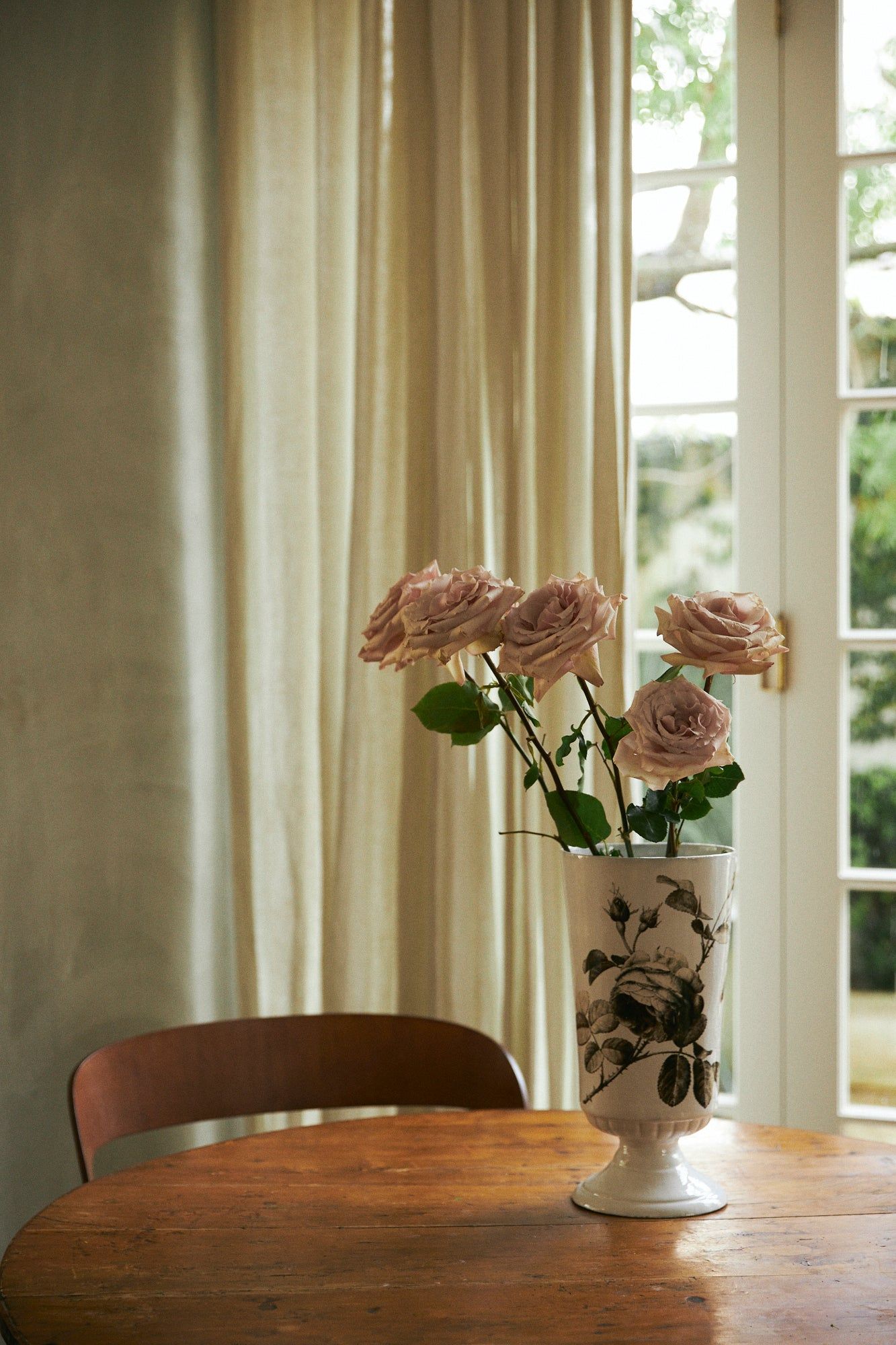 Wooden table with ceramic vase holding pink roses. Beige curtains and glass door let in natural light. Empty chair visible.