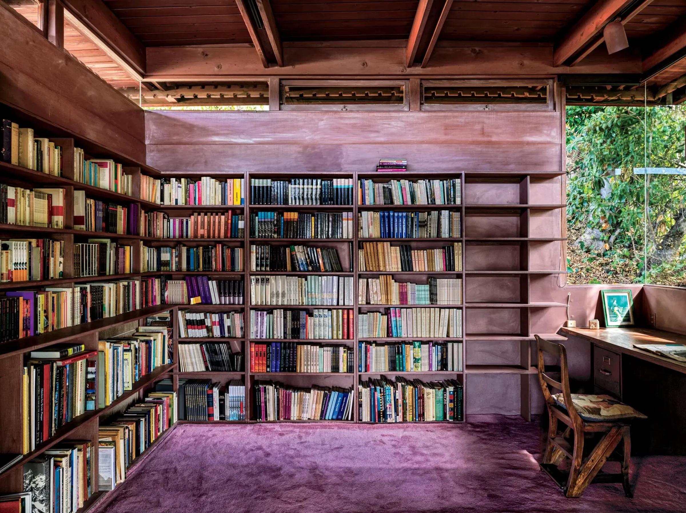 A cozy home library with purple carpet has two walls lined with completely filled bookshelves. A wooden desk and chair by the window look out to greenery outside.
