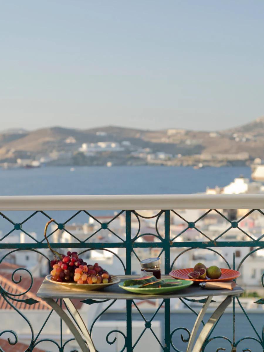 Balcony table set with grapes, figs, and wine. Overlooks coastal town with white buildings, sea, and hills on sunny day.