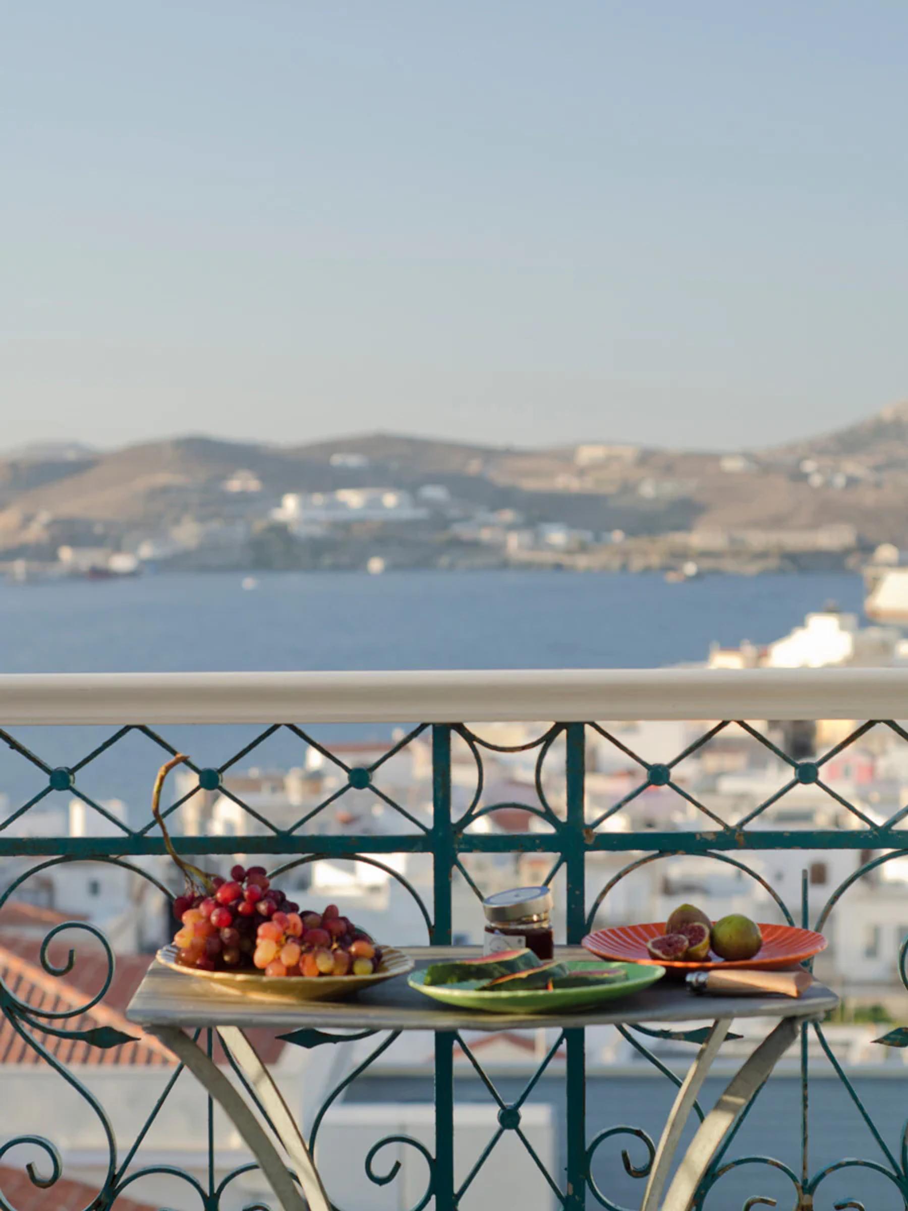 Balcony table set with grapes, figs, and wine. Overlooks coastal town with white buildings, sea, and hills on sunny day.