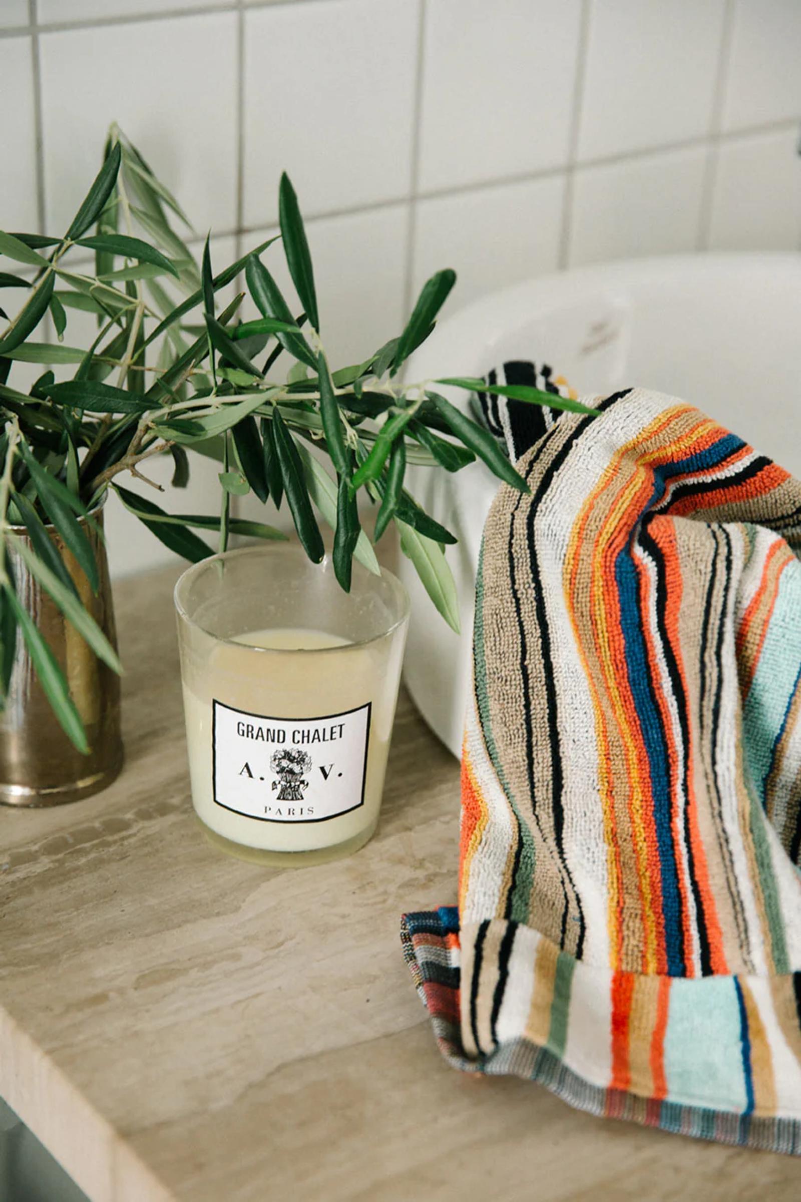 Beige counter with "Grand Chalet Paris" candle and plant. Colourful Missoni-style towel nearby. White tiled wall background.