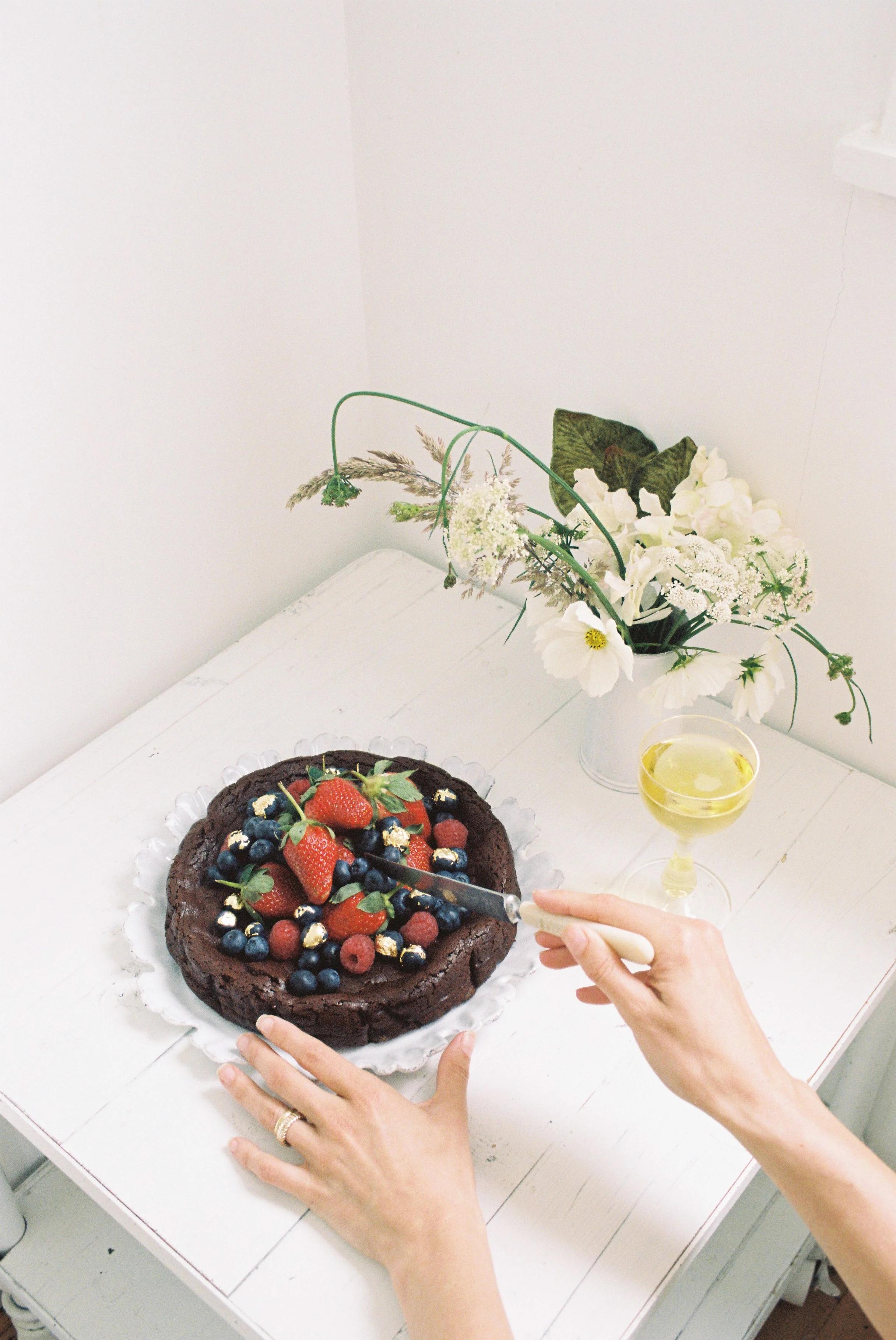 A person cutting a flourless chocolate cake topped with berries and whipped cream, with a glass of wine and a vase of flowers on a white table.