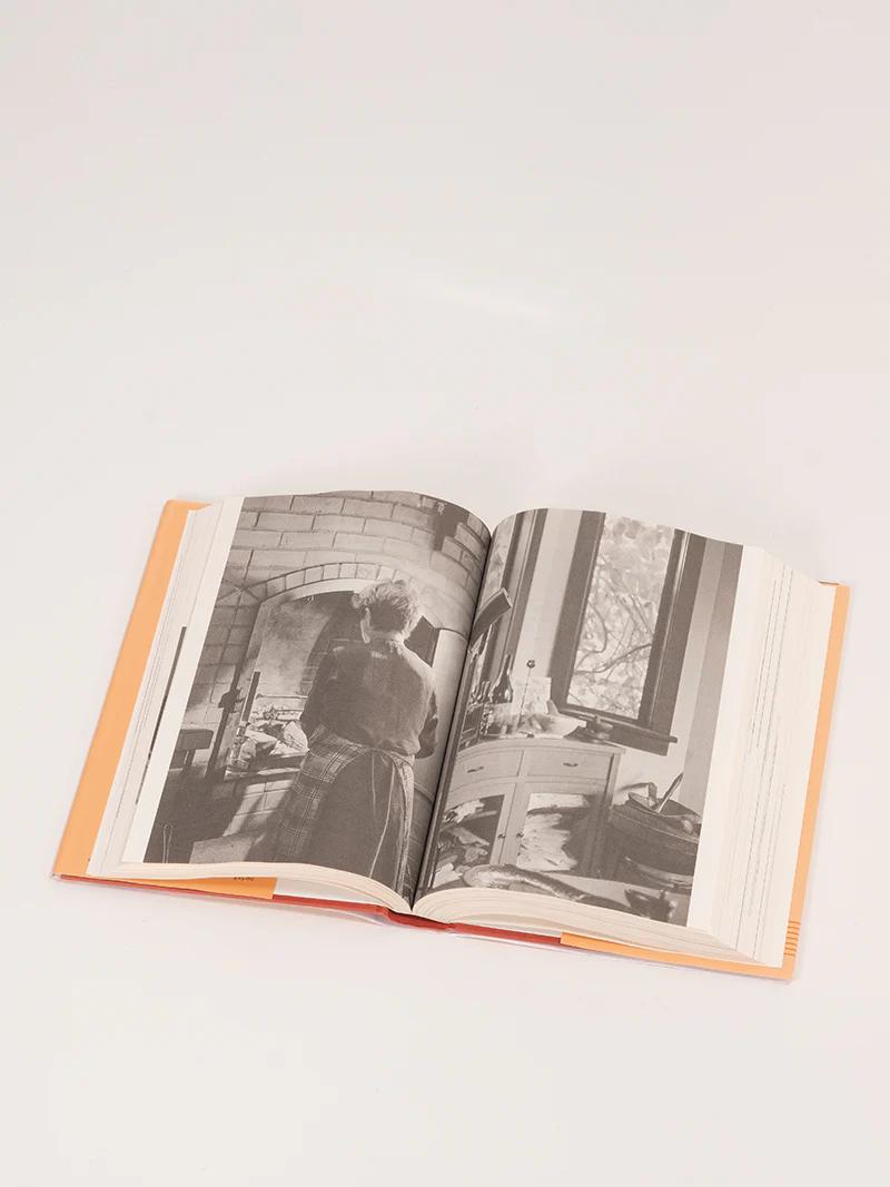 Open book on white surface. Black-and-white photo of person in kitchen visible. Orange book cover.