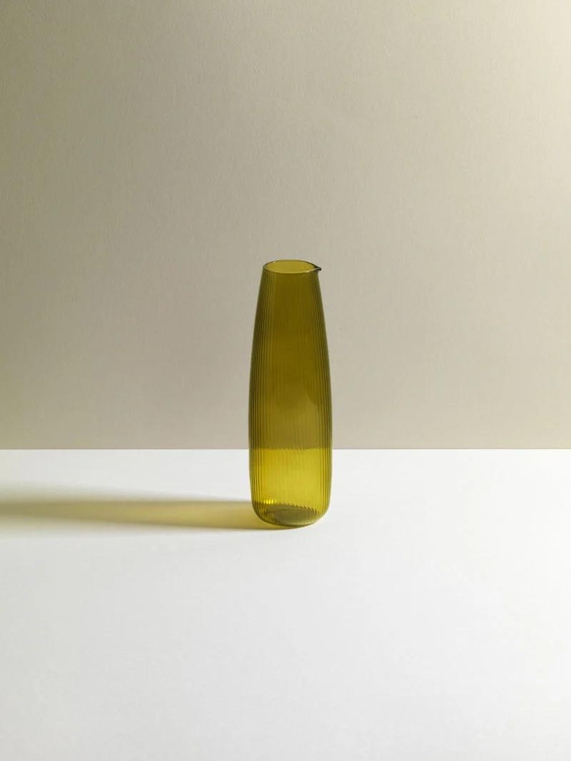 Tall, slender greenish-yellow vase with ribbed texture on smooth surface. Two-toned background creates minimalist look, reflecting Sara Mostofi and Jay Vosoghi's design.