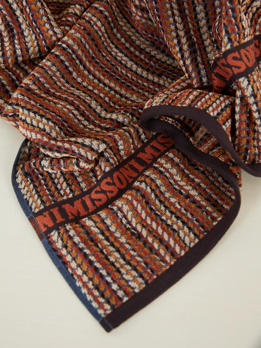 A close-up of a multicoloured houndstooth scarf with "MISSONI" in red. The pattern features brown, orange, beige, and black, showcasing luxurious velour.