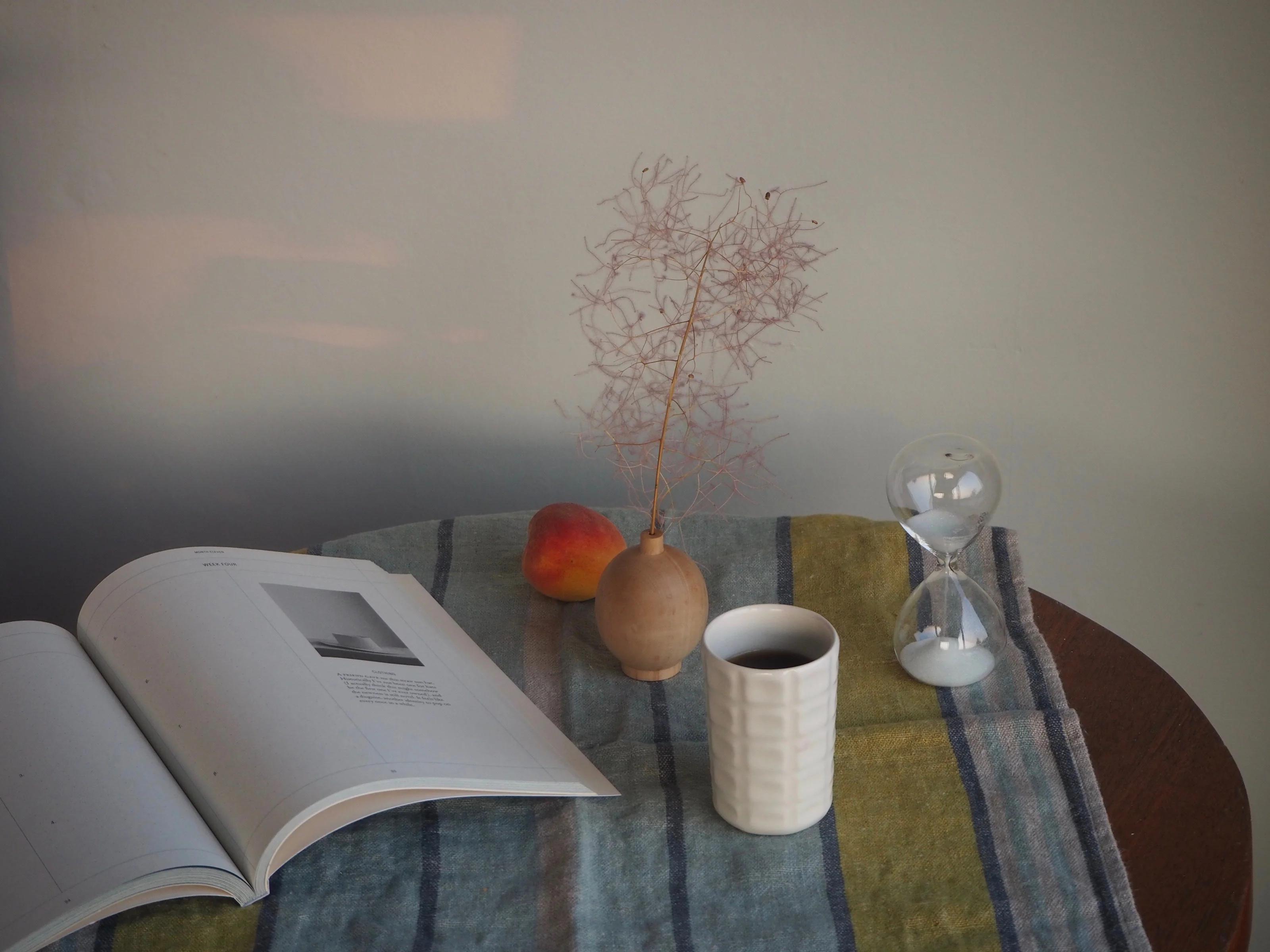 A dawn-lit scene with an open book, a peach, a potted plant, an hourglass, and a coffee cup arranged on a striped cloth.