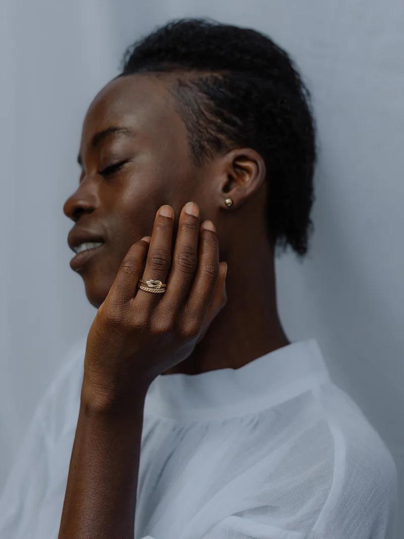 Profile of person with short hair, closed eyes, white top. Touching face, wearing gemstone ring and small earring.