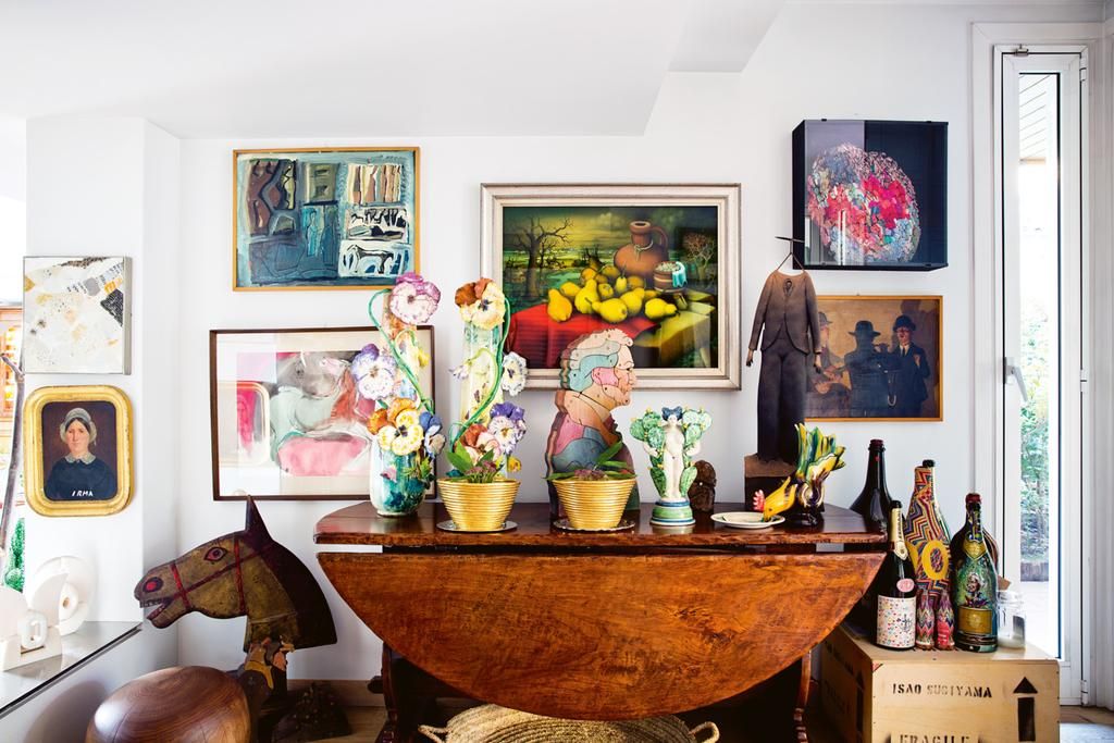 Vibrant living room with wooden table, ceramic flowers, and plants. Eclectic artworks on walls. Vintage rocking horse and decorative bottles.