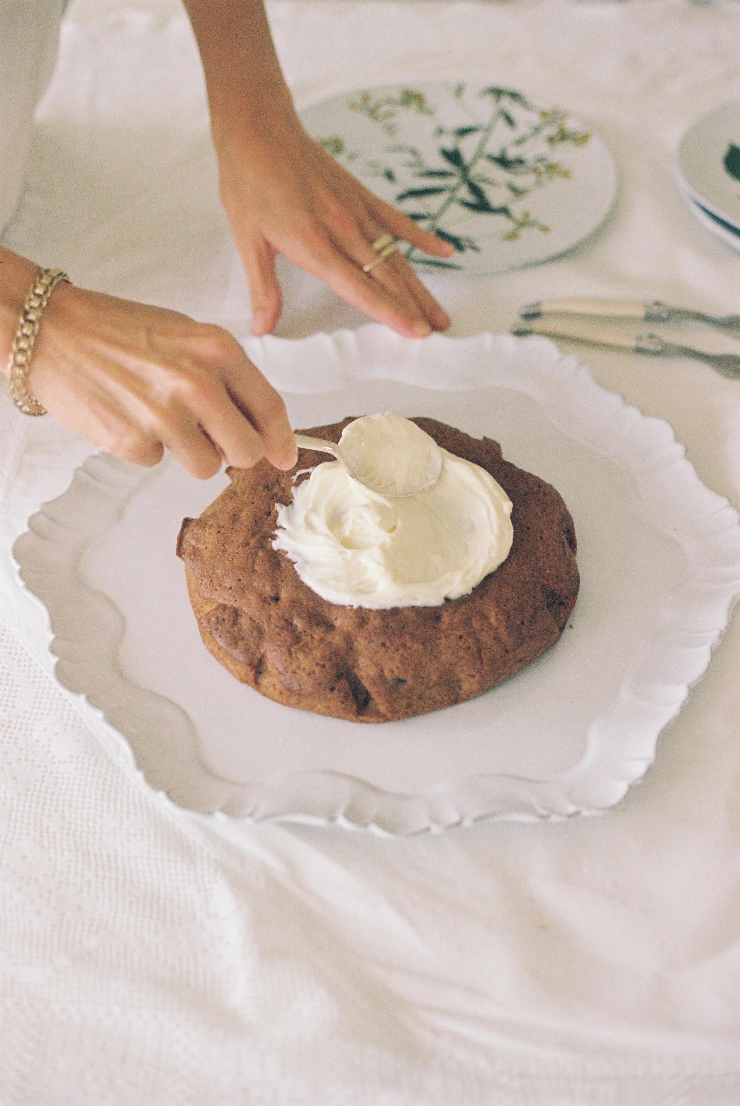 Person frosting brown carrot cake on scalloped platter. White tablecloth, patterned plate and utensils in background.