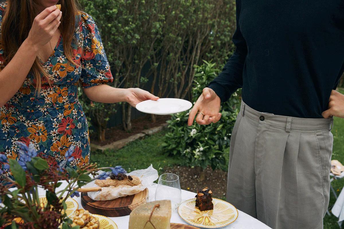 Two people at food-laden table. One holding white plate, other reaching to take it.