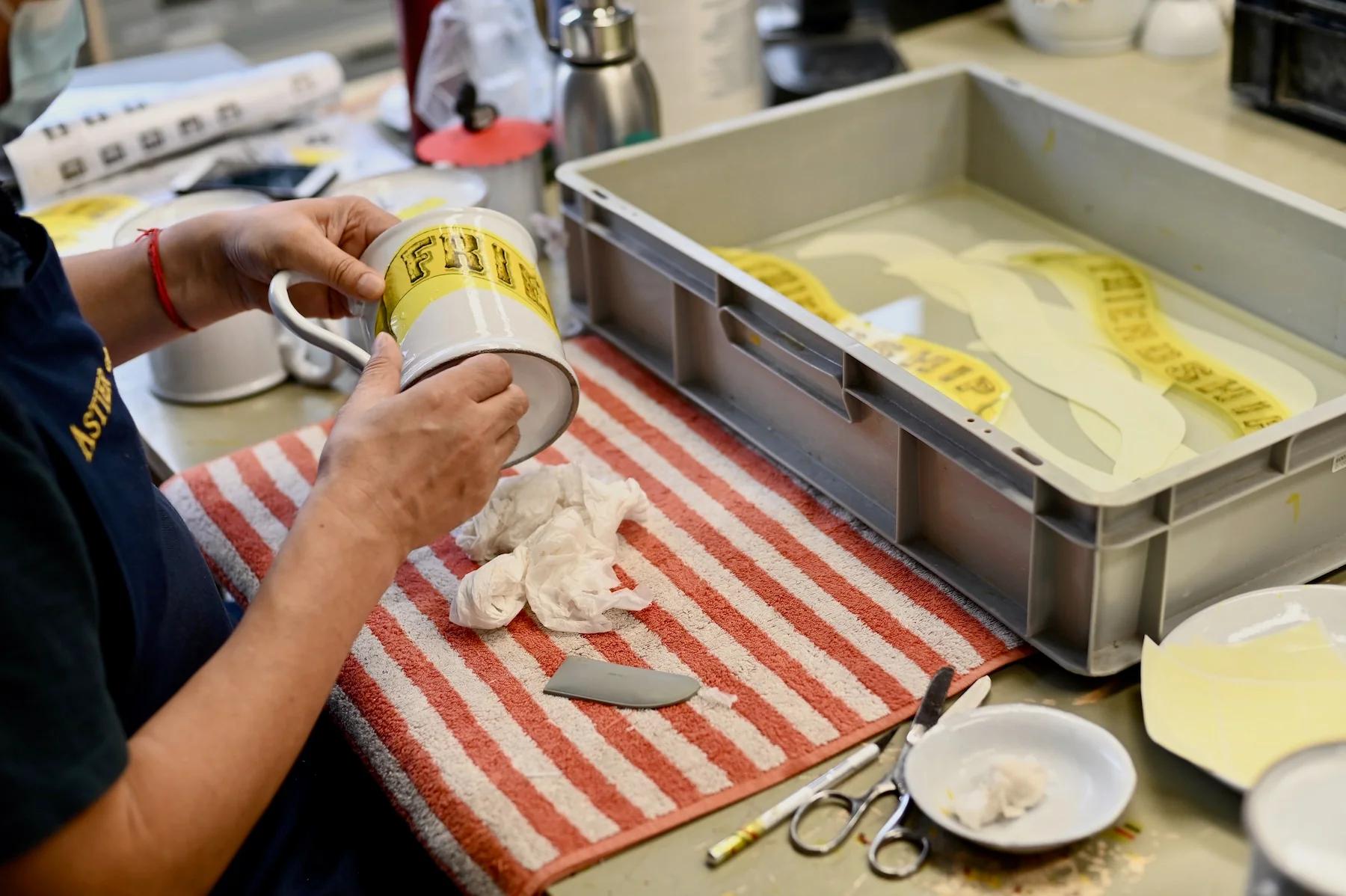 Person applying decal to white mug. Water trays, scissors, and yellow decals on striped towel nearby. Person's shirt displays text, face unseen.