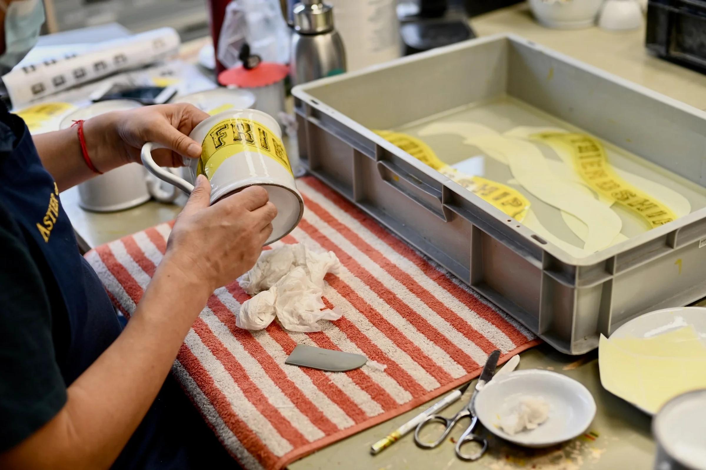 Person applying decal to white mug. Water trays, scissors, and yellow decals on striped towel nearby. Person's shirt displays text, face unseen.