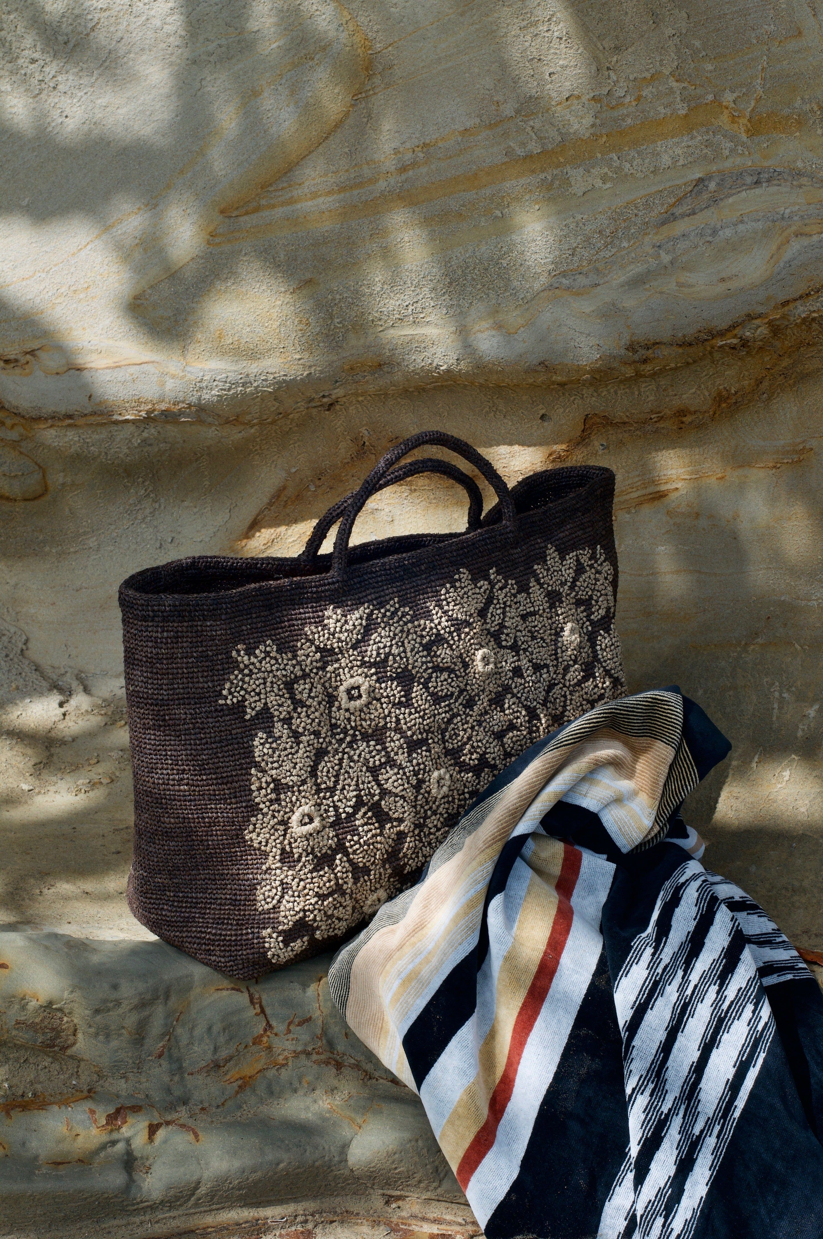 Woven handbag with floral patterns on textured wall. Multicoloured striped fabric draped nearby. Leaf shadows visible.