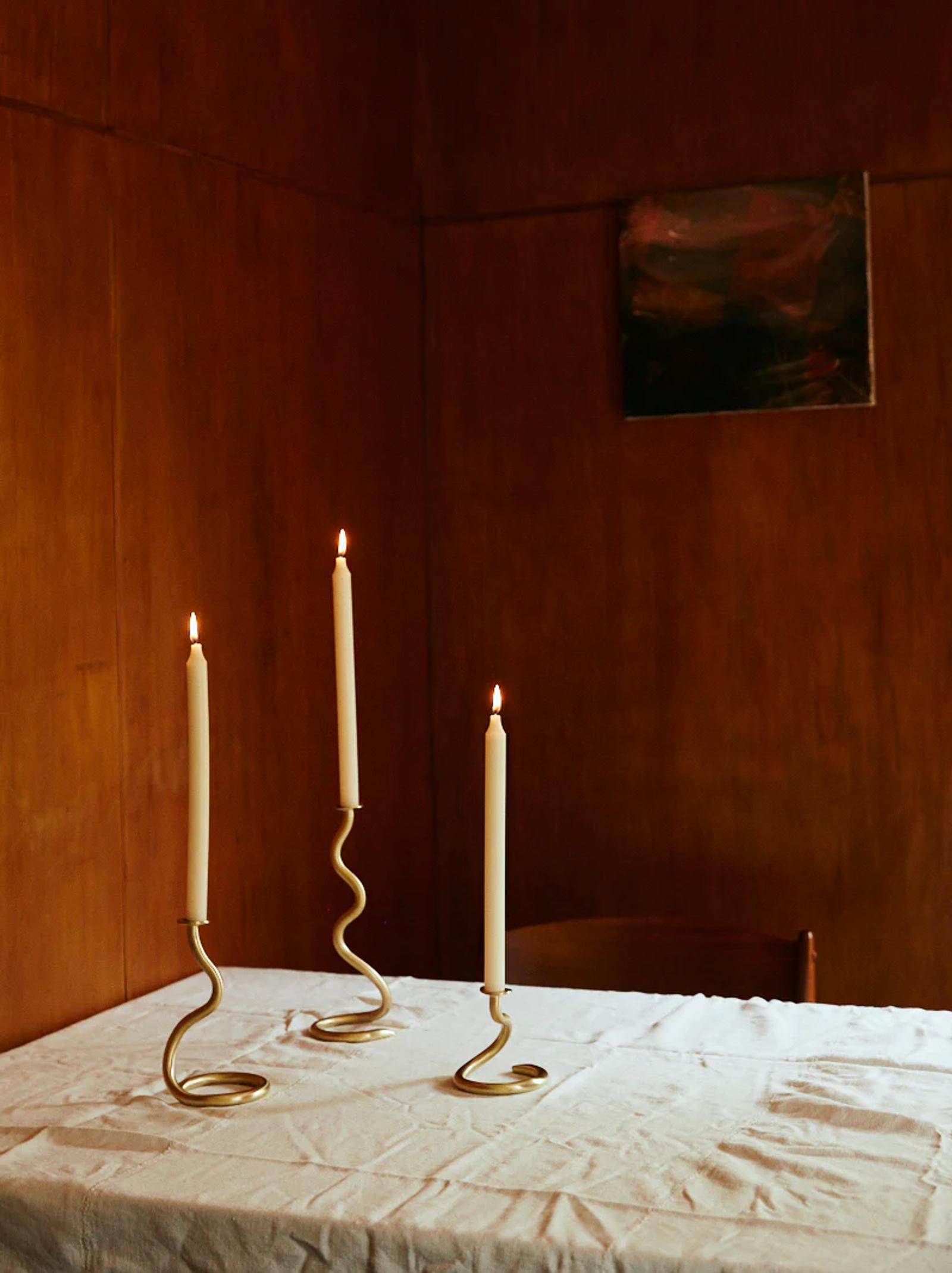 Three lit white candles on gold holder on white tablecloth. Wooden-panelled wall with framed abstract painting behind.