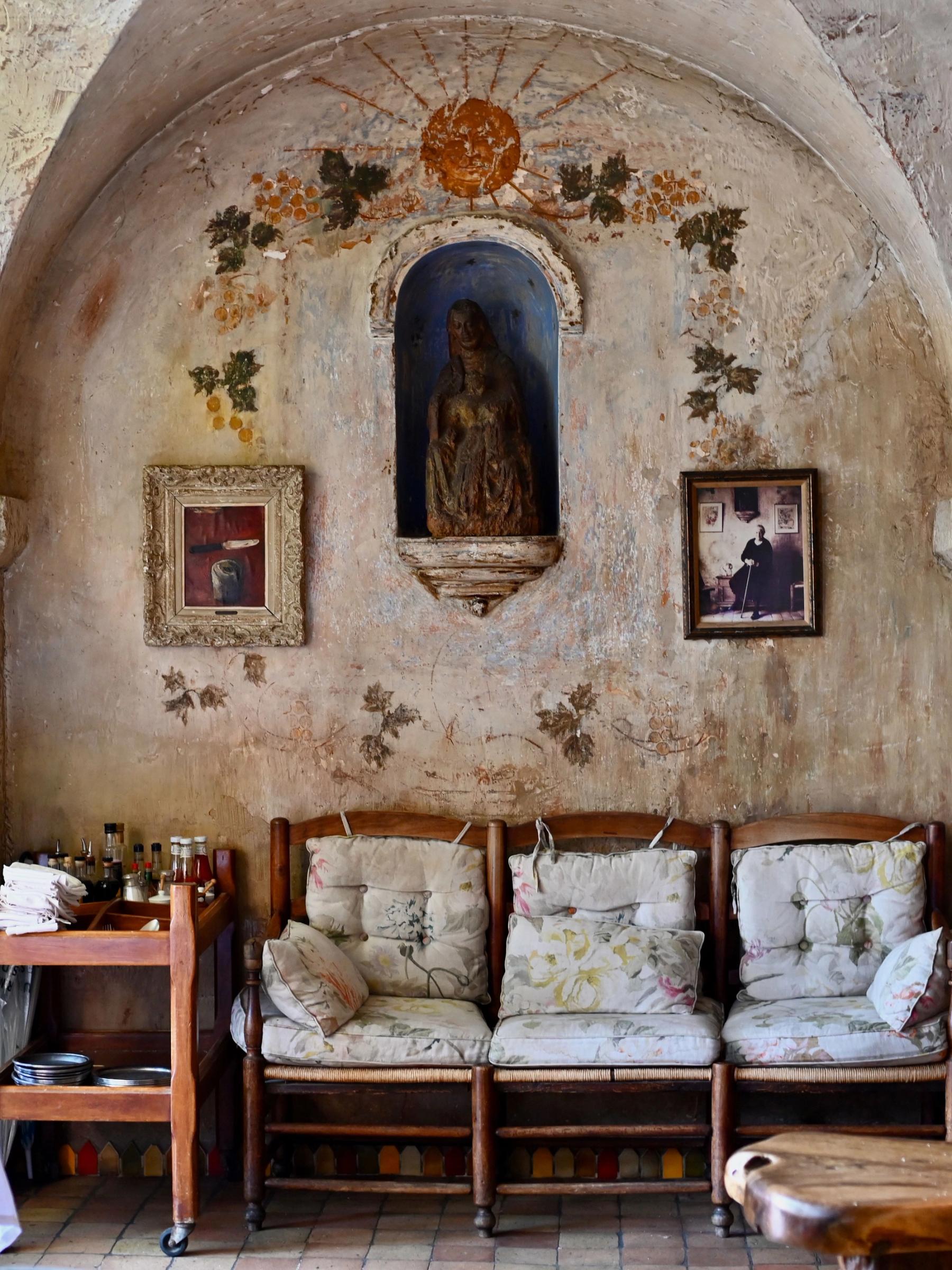 Rustic seating area with arched ceilings and stone alcove housing a small statue. Framed pictures and ivy adorn walls, with wooden chairs nearby.