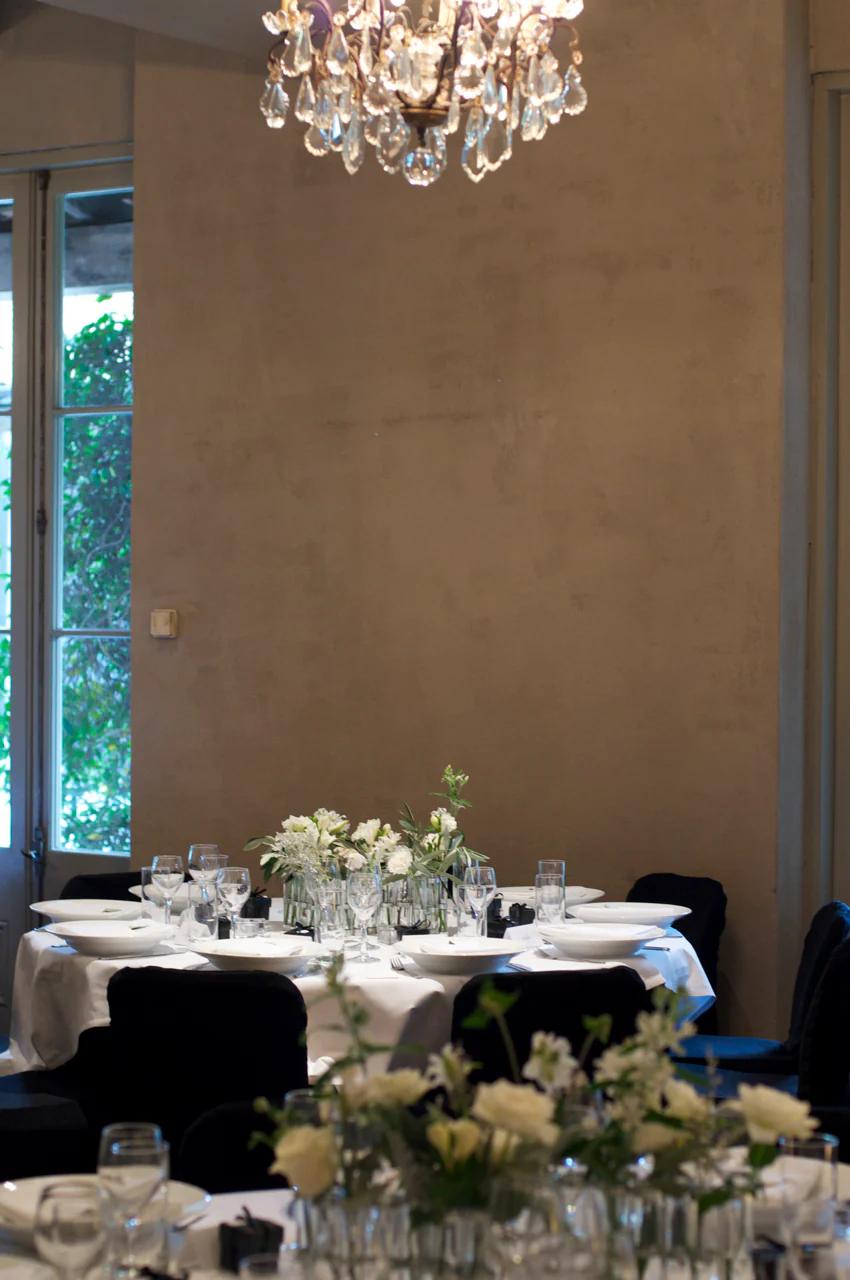 Round table set for meal with white plates, wine glasses, and floral centrepieces. Large window and chandelier in background.