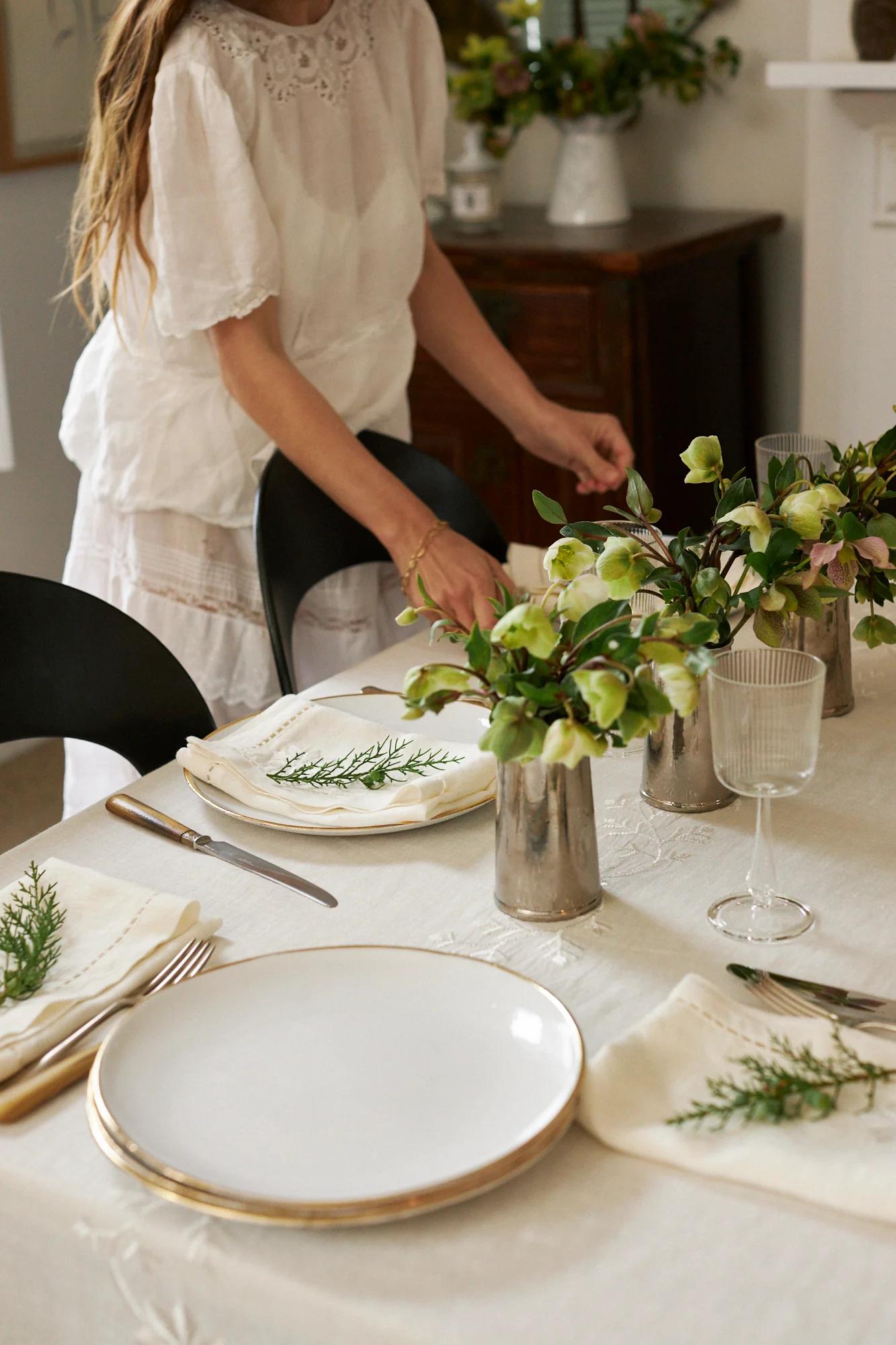 A person in white arranges a table with plates, napkins, and floral centrepieces. The embroidered tablecloth and backdrop create a festive atmosphere.