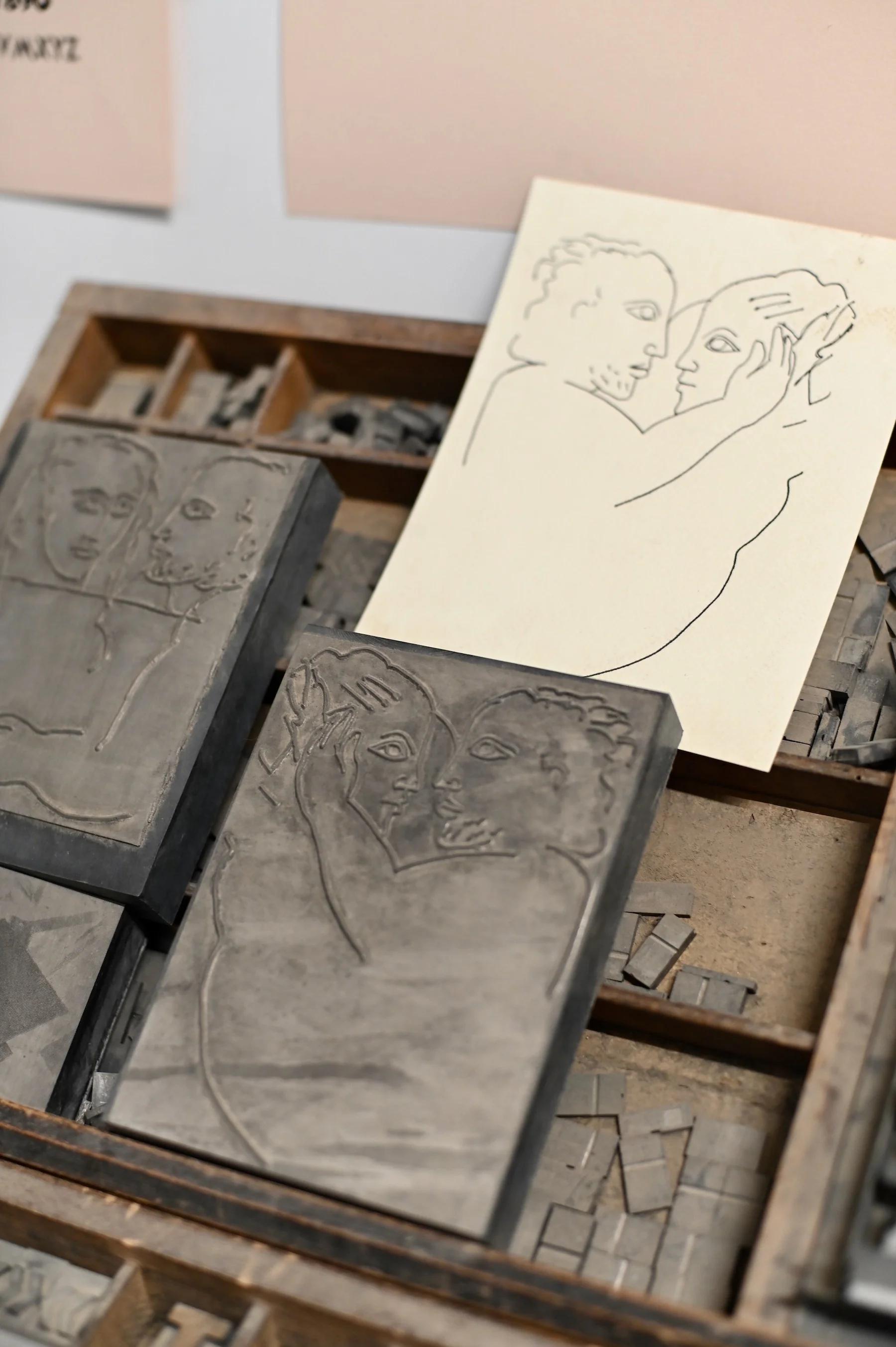 Tabletop with engraved stone blocks depicting entwined figures. Matching sketch on paper. Wooden compartments with small metal pieces surrounding.