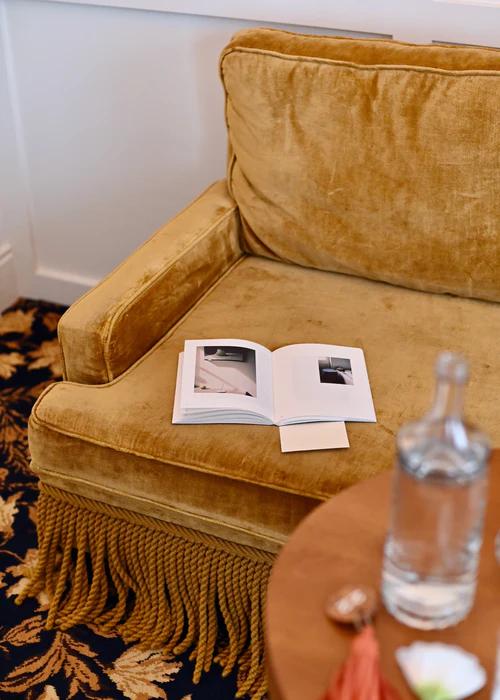 A mustard yellow velvet armchair with a long floor fringe trim sits on a floral-patterned carpet. An open book lies on the seat. In the foreground, a partially visible round wooden table holds a clear water bottle and a blurred small object from Tessuti's Journal homeware artisans collection.