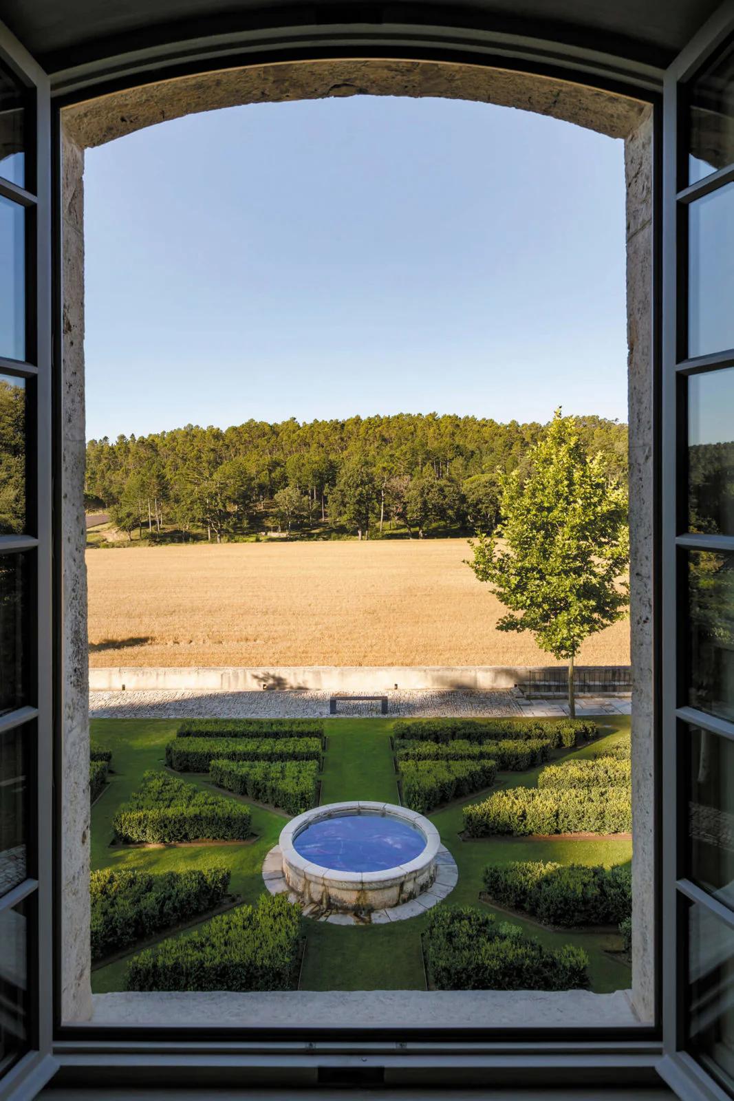 A château window view of a landscaped garden with a fountain. Beyond lie golden fields and a dense forest under a clear blue sky.