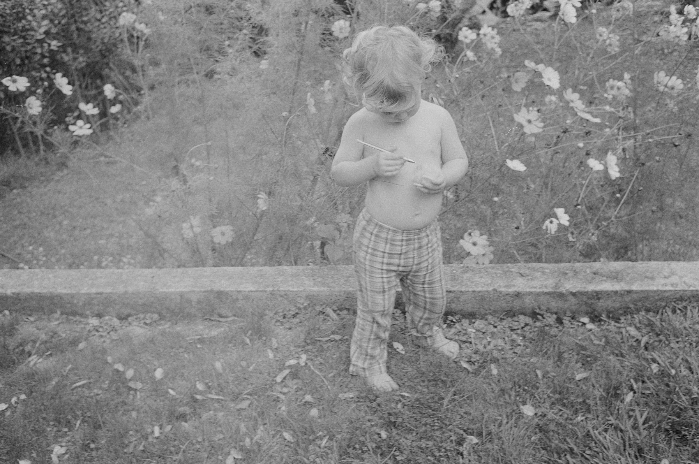 Black and white photo of barefoot toddler in plaid pants. Holding object, standing in grassy area with flowers.