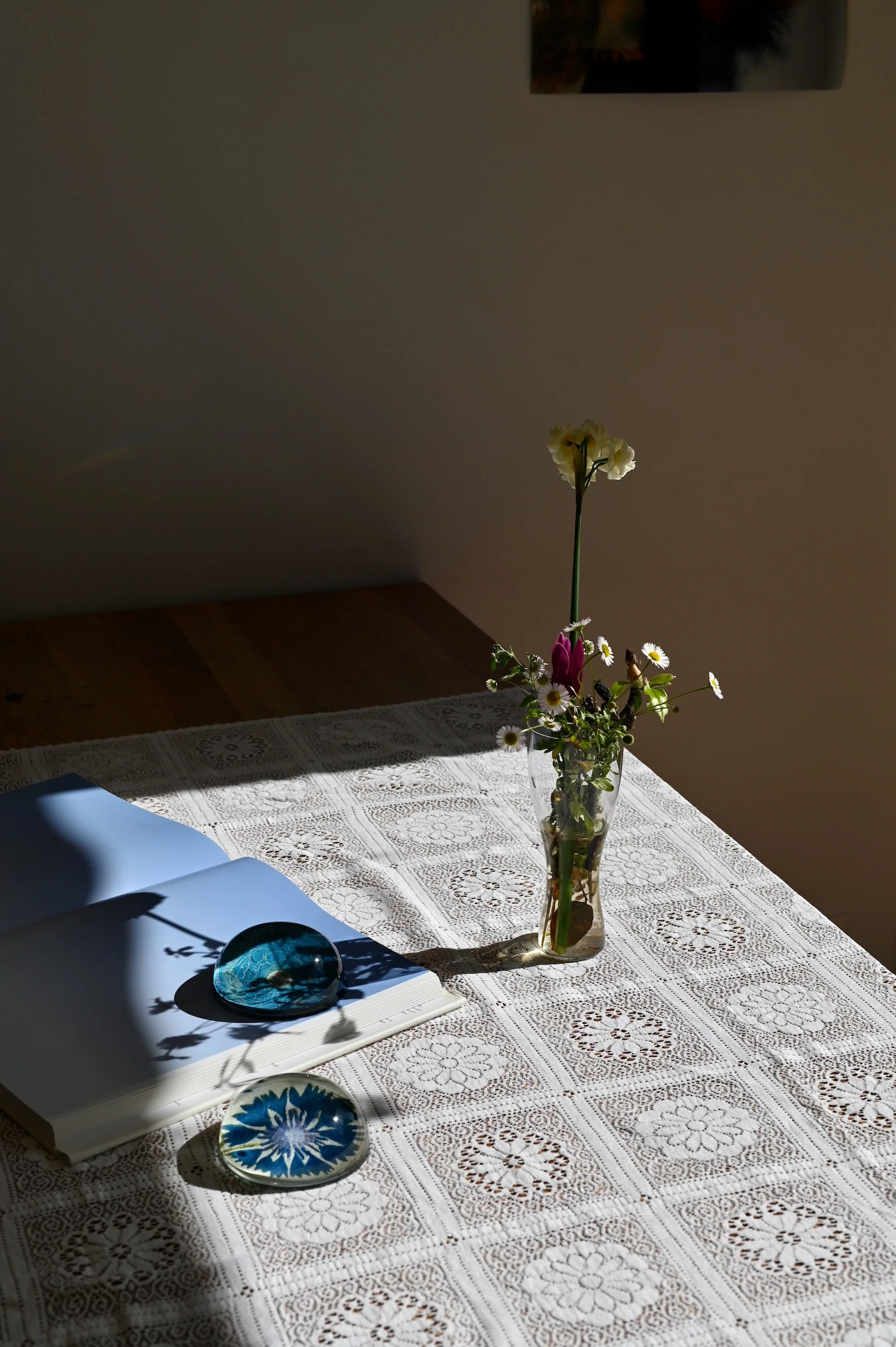 Vase with yellow and white flowers on lace tablecloth. Open book, blue glass paperweight. Sunlit scene.