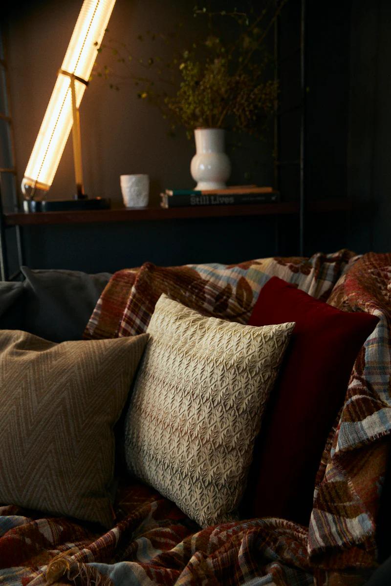 Couch with patterned pillows and plaid blanket. Tall lamp near shelf with vase and books. Warm, inviting scene with rich textures.