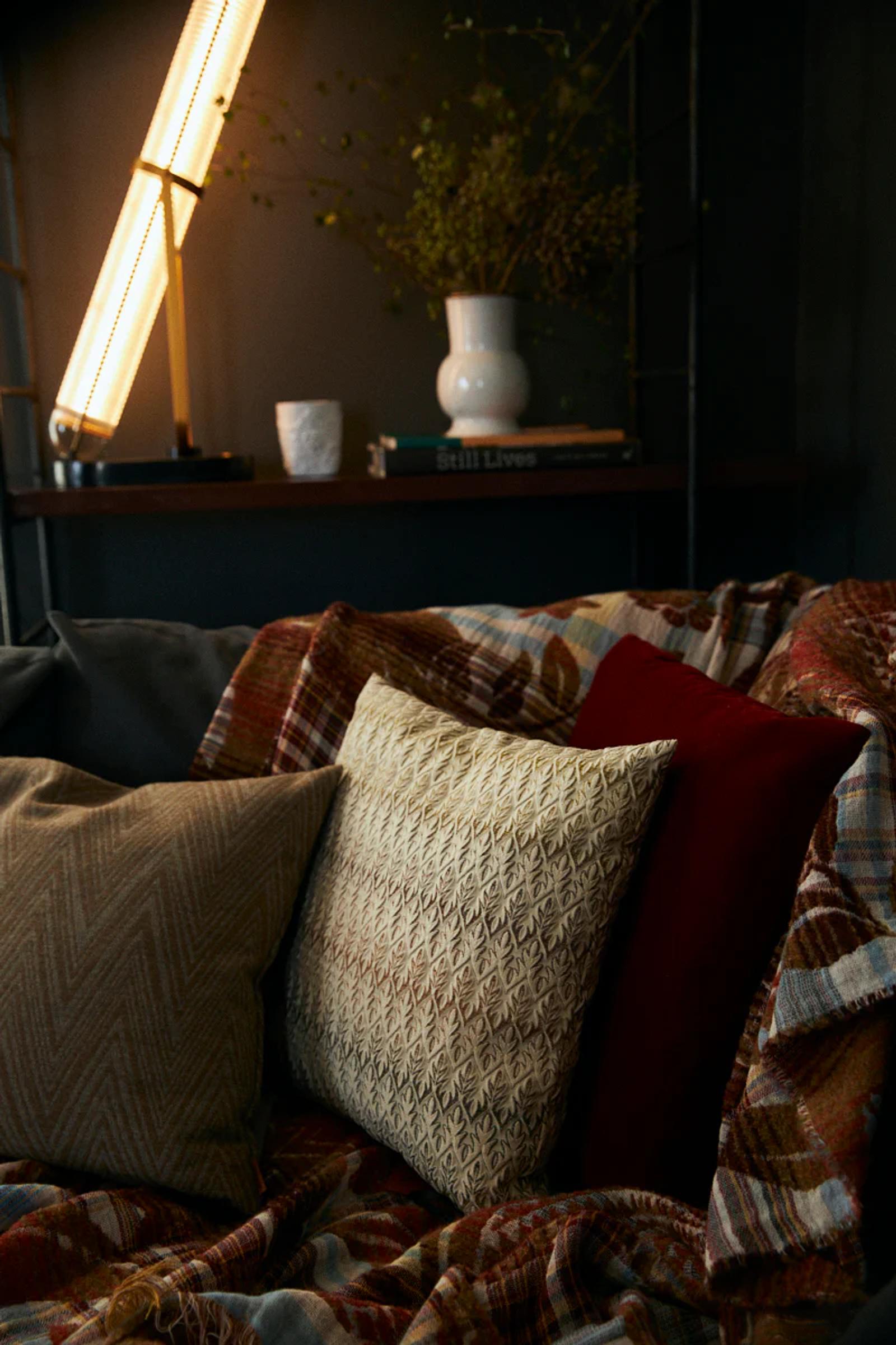 Couch with patterned pillows and plaid blanket. Tall lamp near shelf with vase and books. Warm, inviting scene with rich textures.