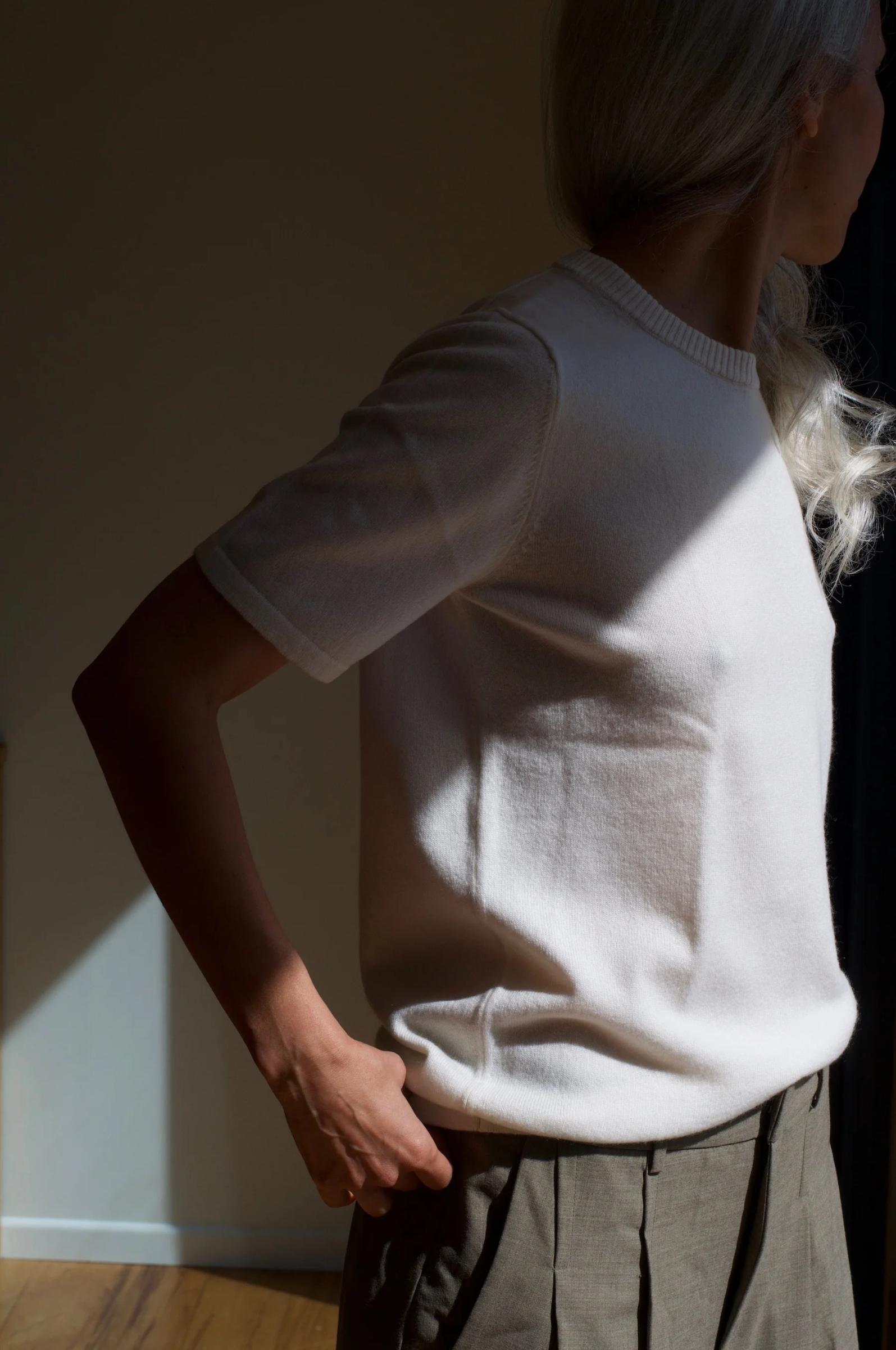 Long-haired person in sunlit area wearing light knit top and high-waisted trousers. Hand on hip, partially turned away.
