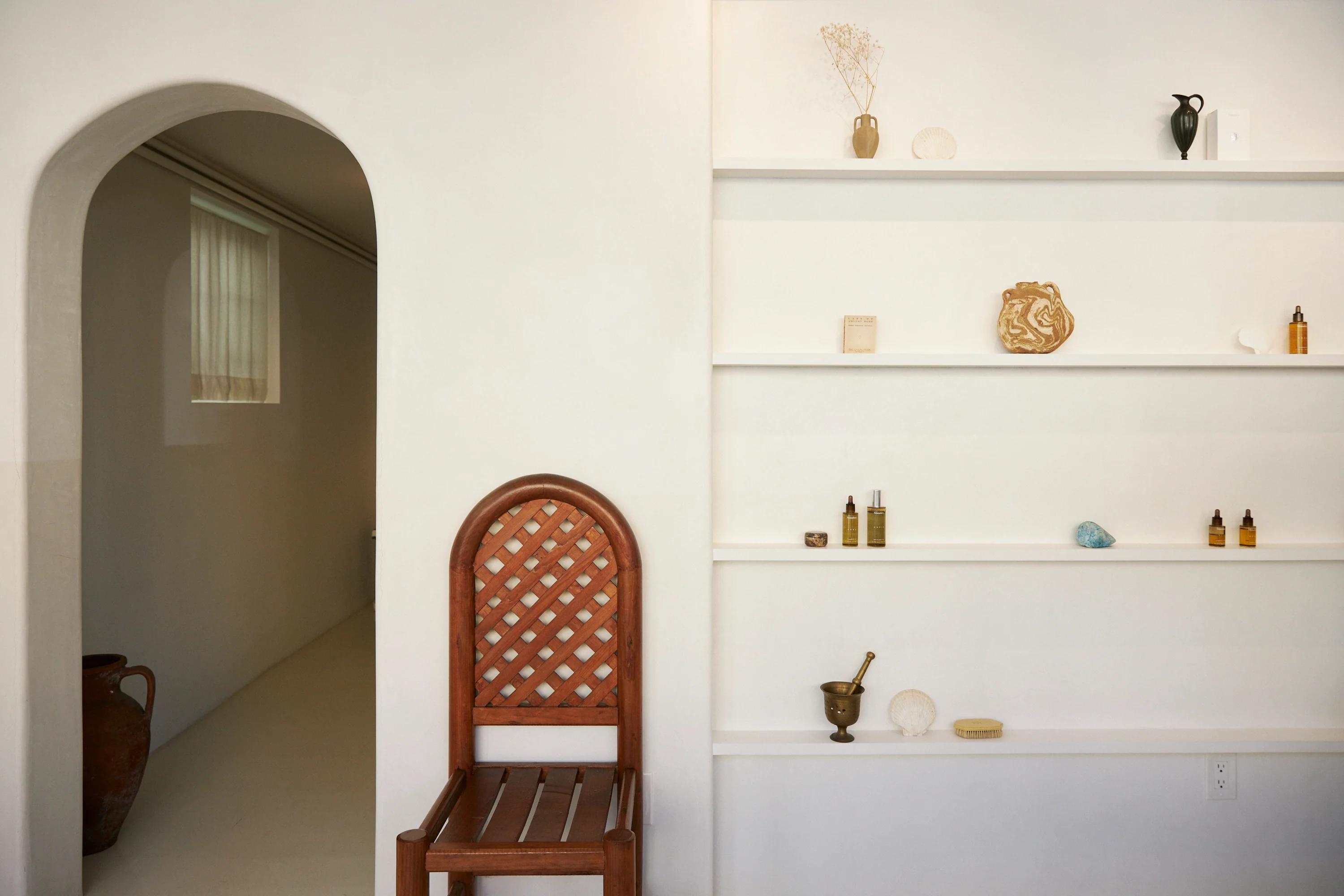 Wooden chair by white wall with shelves. Shelves hold decor items, skincare products, and dried flowers. Arched doorway to sunlit hallway.