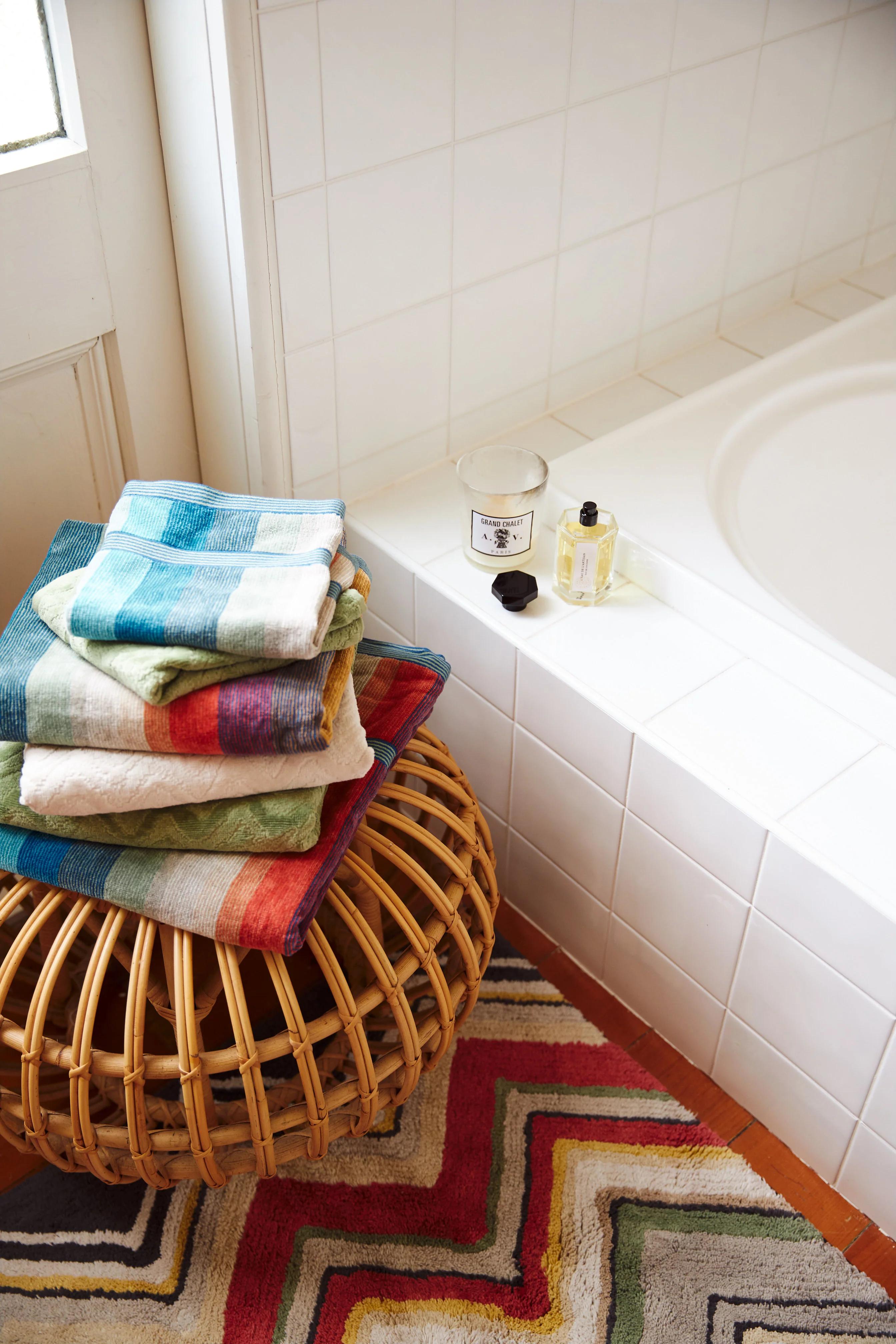 Colourful towels on wicker stool by bathtub. Candle and bottle on tub edge. Zigzag-patterned rug on floor.