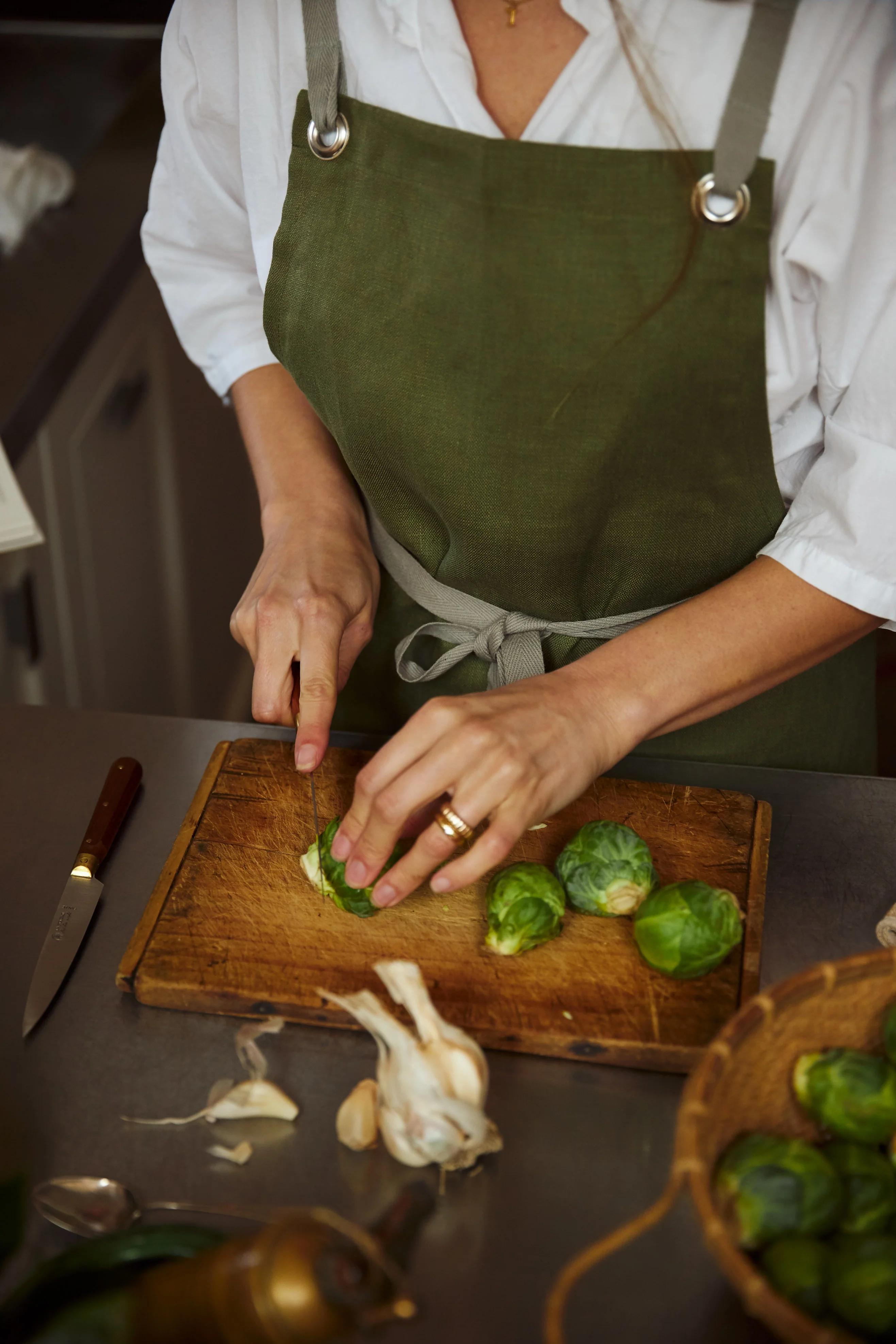 Person in green apron chopping Brussels sprouts. Garlic, knife, and vegetable basket on countertop.