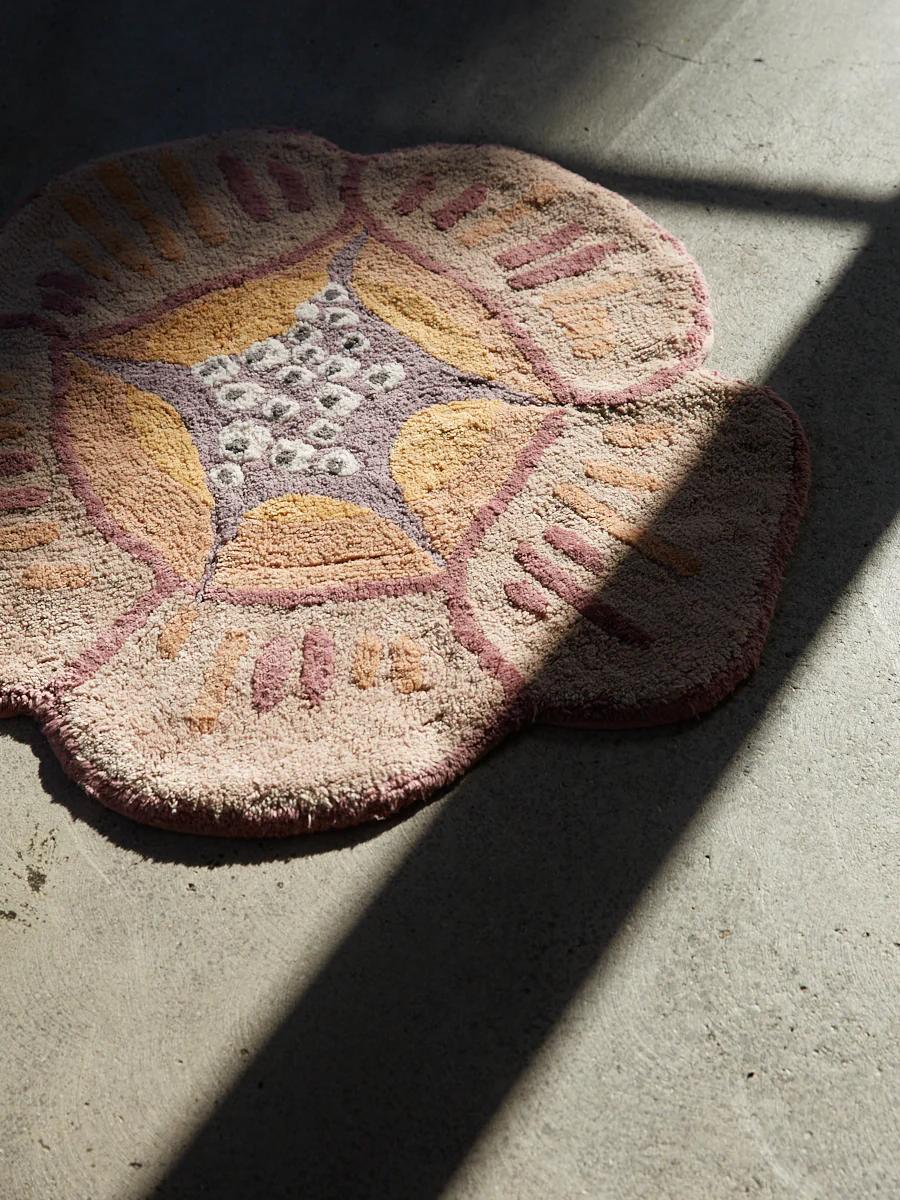 A flower-shaped bath mat in pink, yellow, and purple on a grey concrete floor, with sunlight casting diagonal shadows.