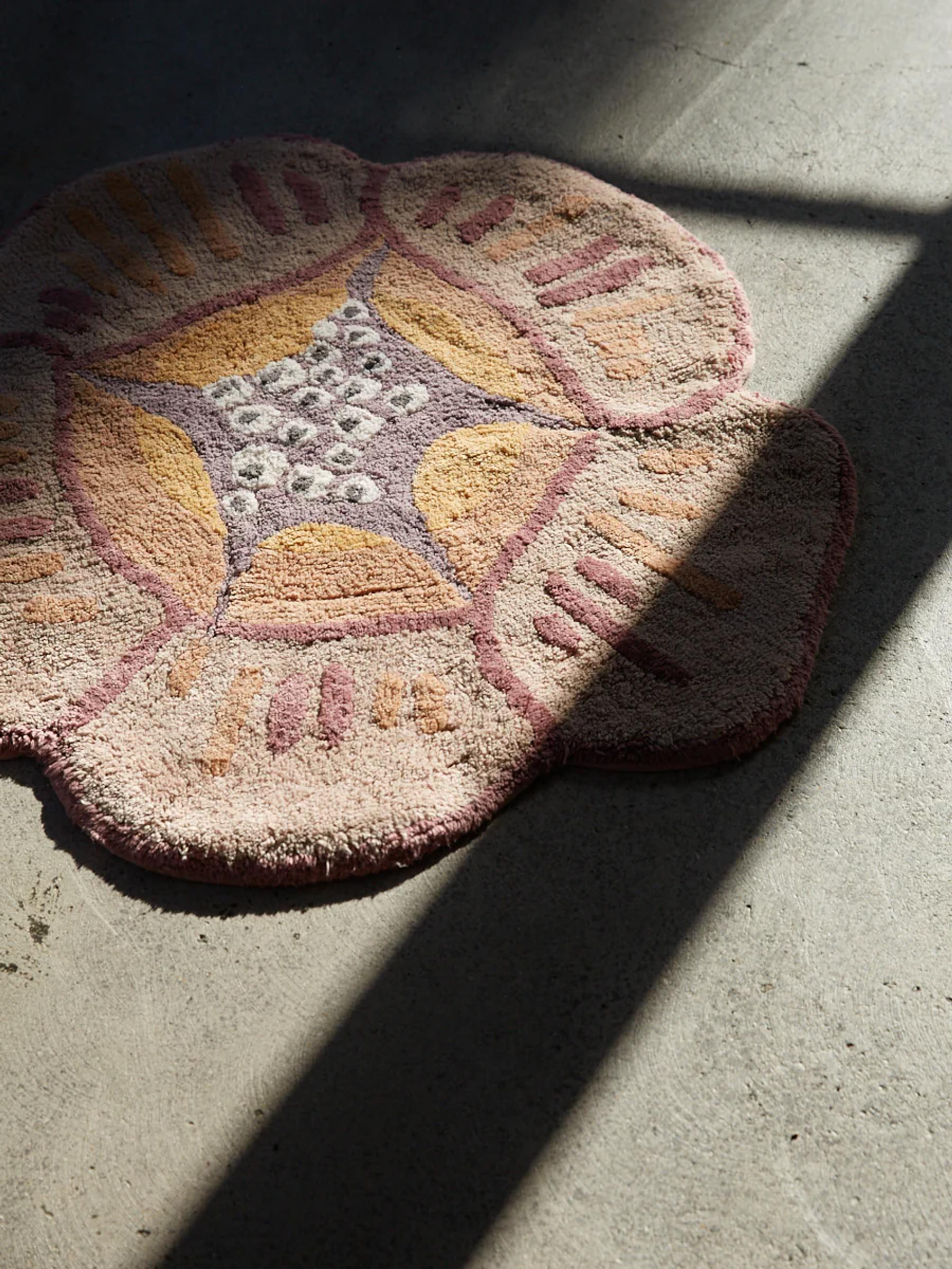 A flower-shaped bath mat in pink, yellow, and purple on a grey concrete floor, with sunlight casting diagonal shadows.