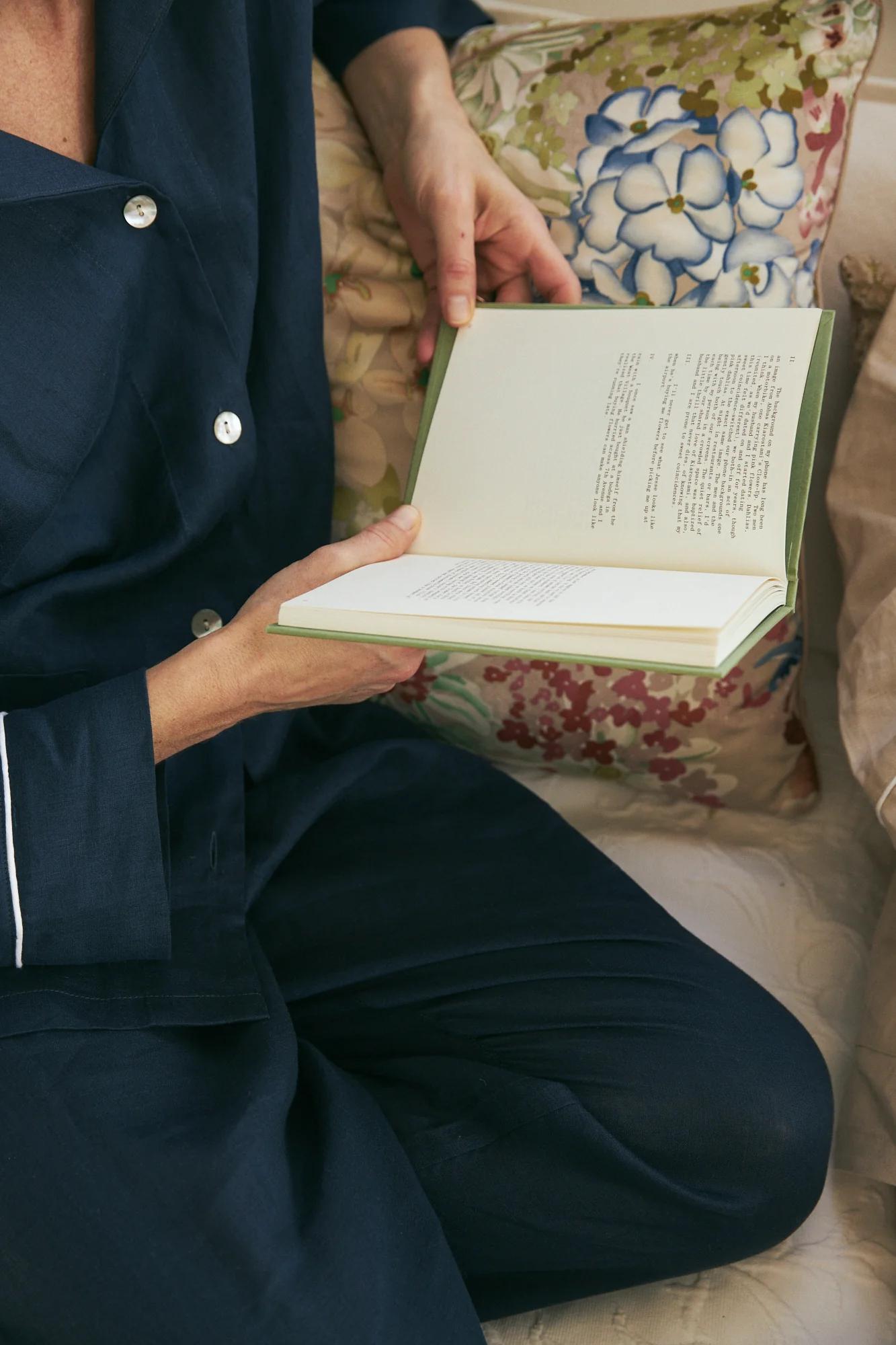 Person in dark blue pyjamas reading book while sitting on floral cushion. Cosy, comfortable setting.