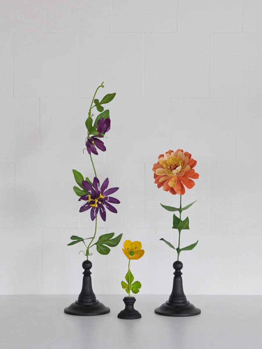 Three flowers in black vases: purple, yellow, and orange blooms. Light grey background.