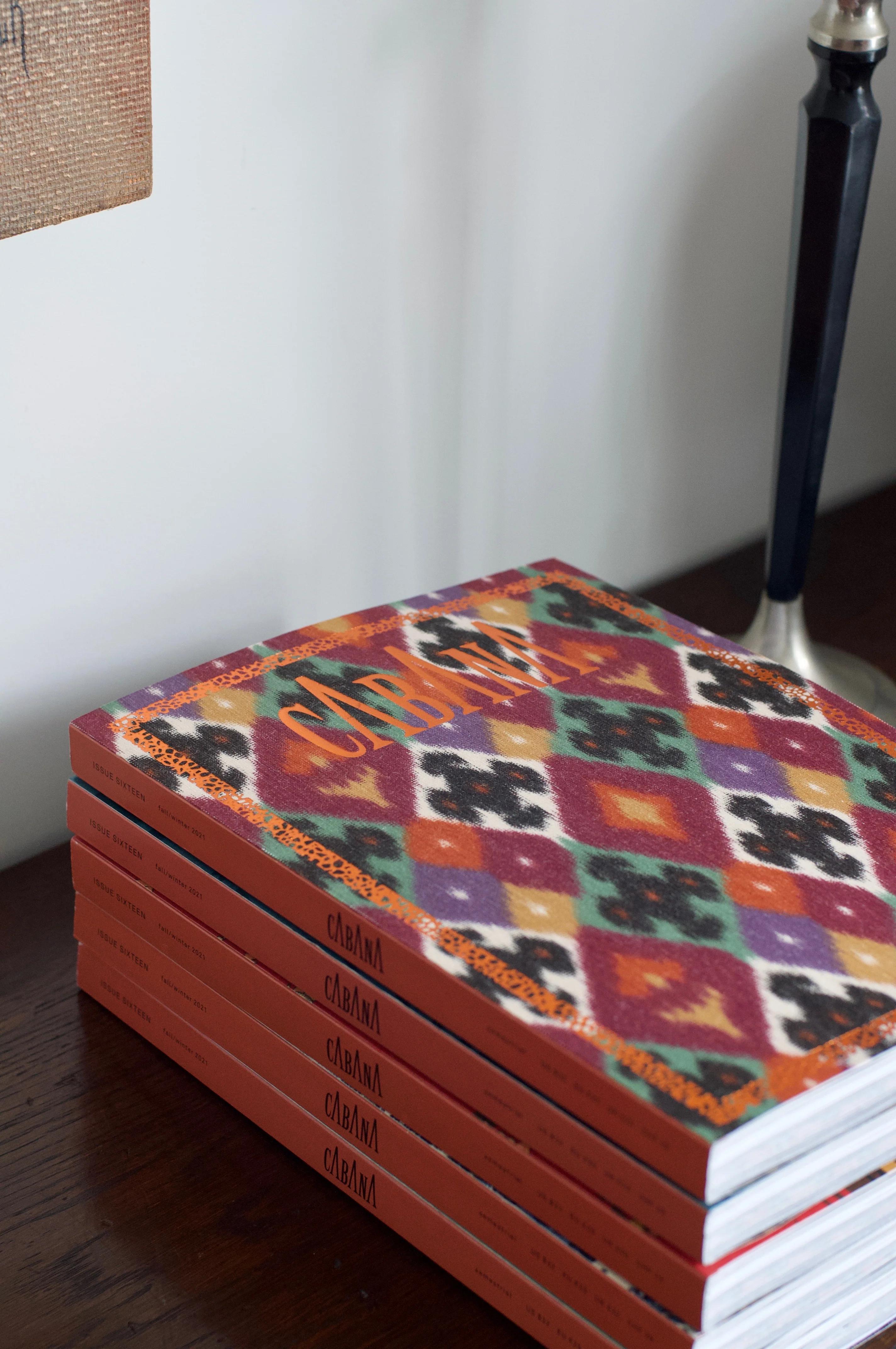 Five CABANA Magazines stacked on wood. Top issue has colourful geometric pattern. Lamp base visible.