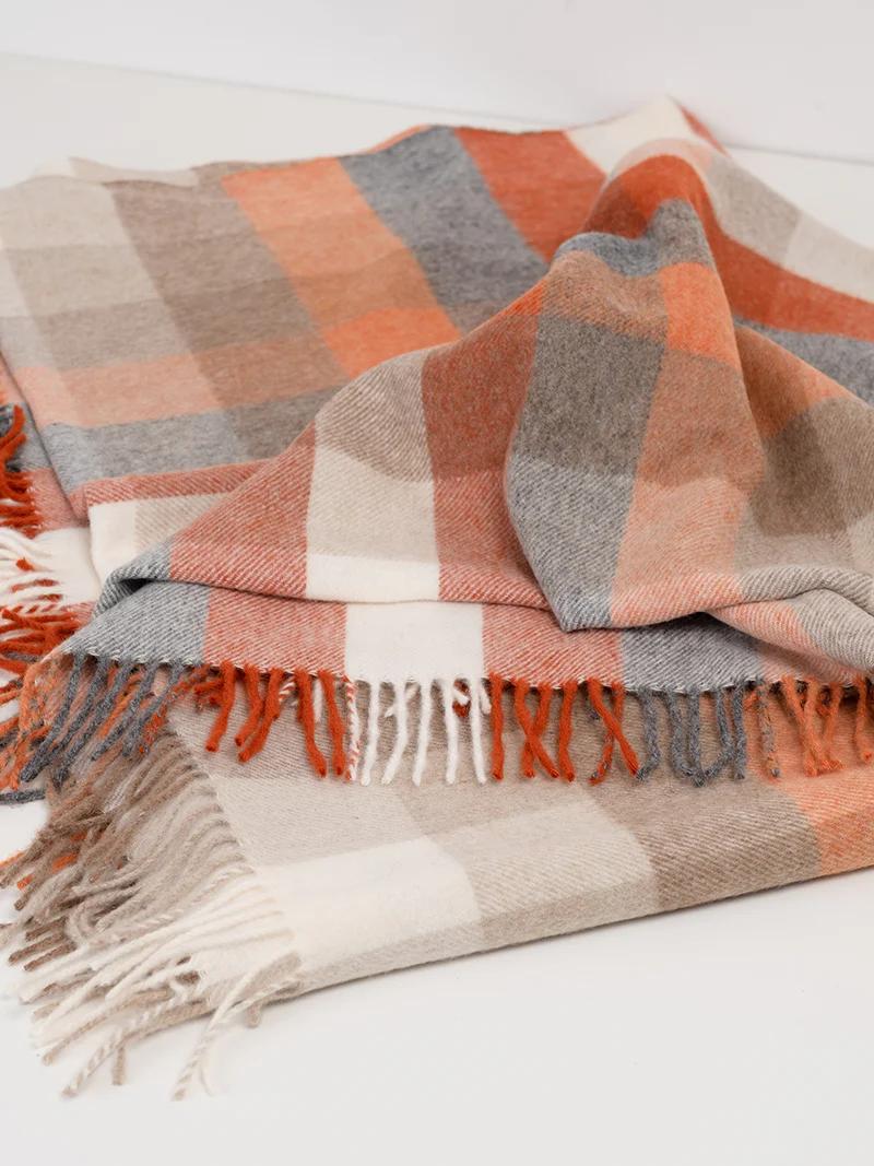 Folded blanket with plaid pattern in orange, beige, grey, and white. The blanket’s fringed edges and cozy texture are highlighted by a creatively folded top layer.