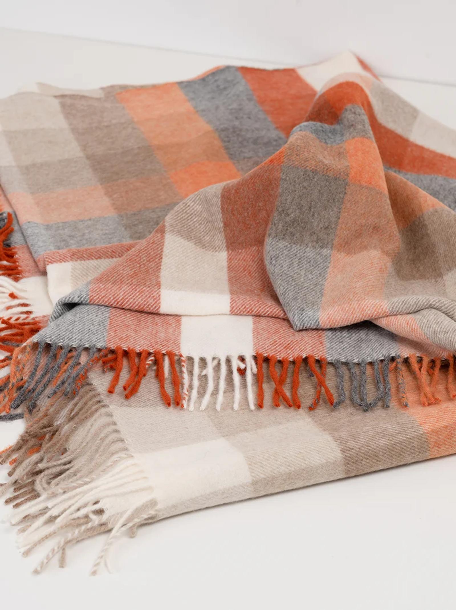 Folded blanket with plaid pattern in orange, beige, grey, and white. The blanket’s fringed edges and cozy texture are highlighted by a creatively folded top layer.