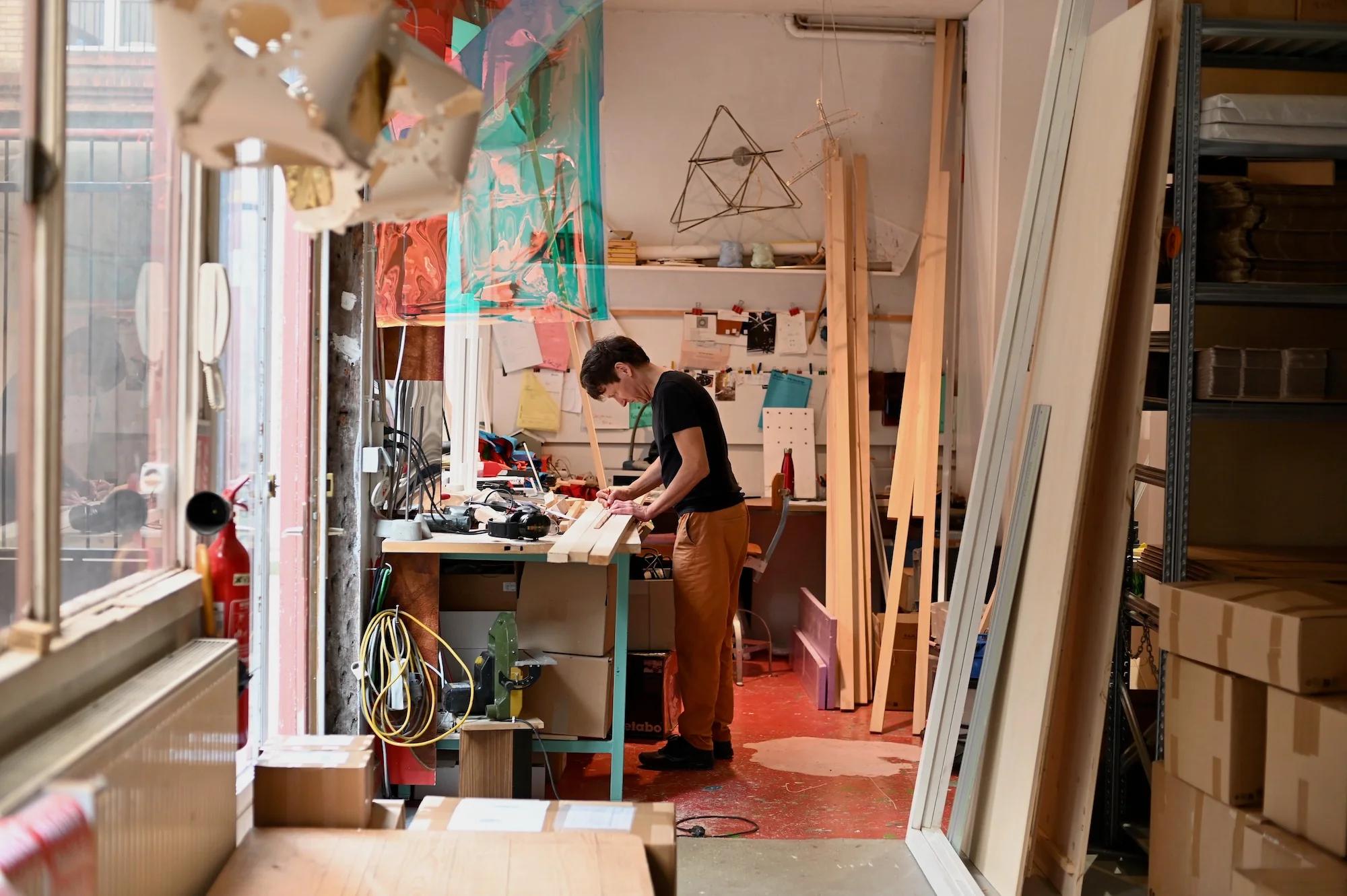 Person in black shirt, orange pants at cluttered workbench. Well-lit woodshop with tools, planks, shelves. Large window.