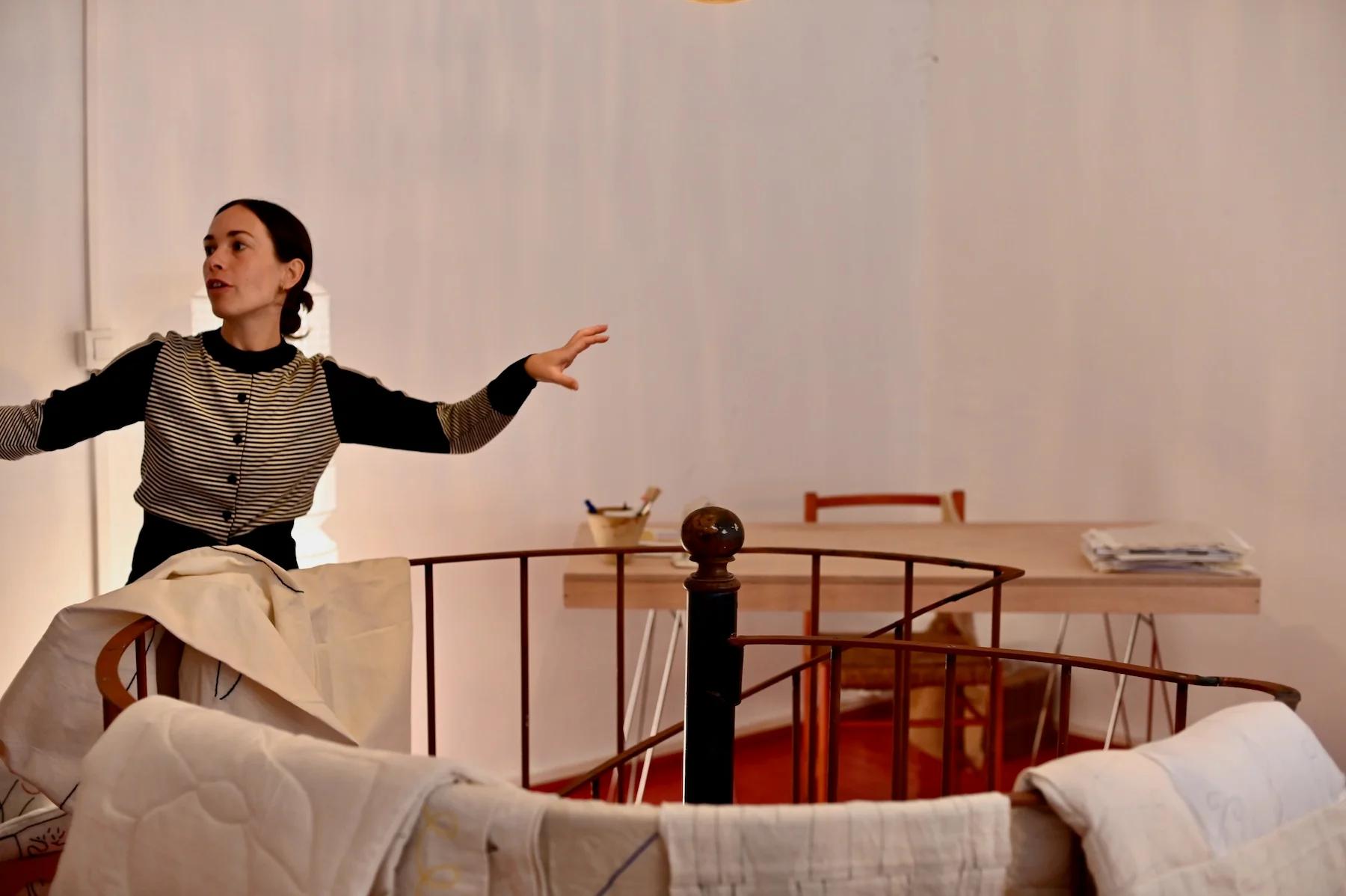 Woman with dark hair gestures near spiral staircase. Wooden table and chairs with linens in minimally furnished room.