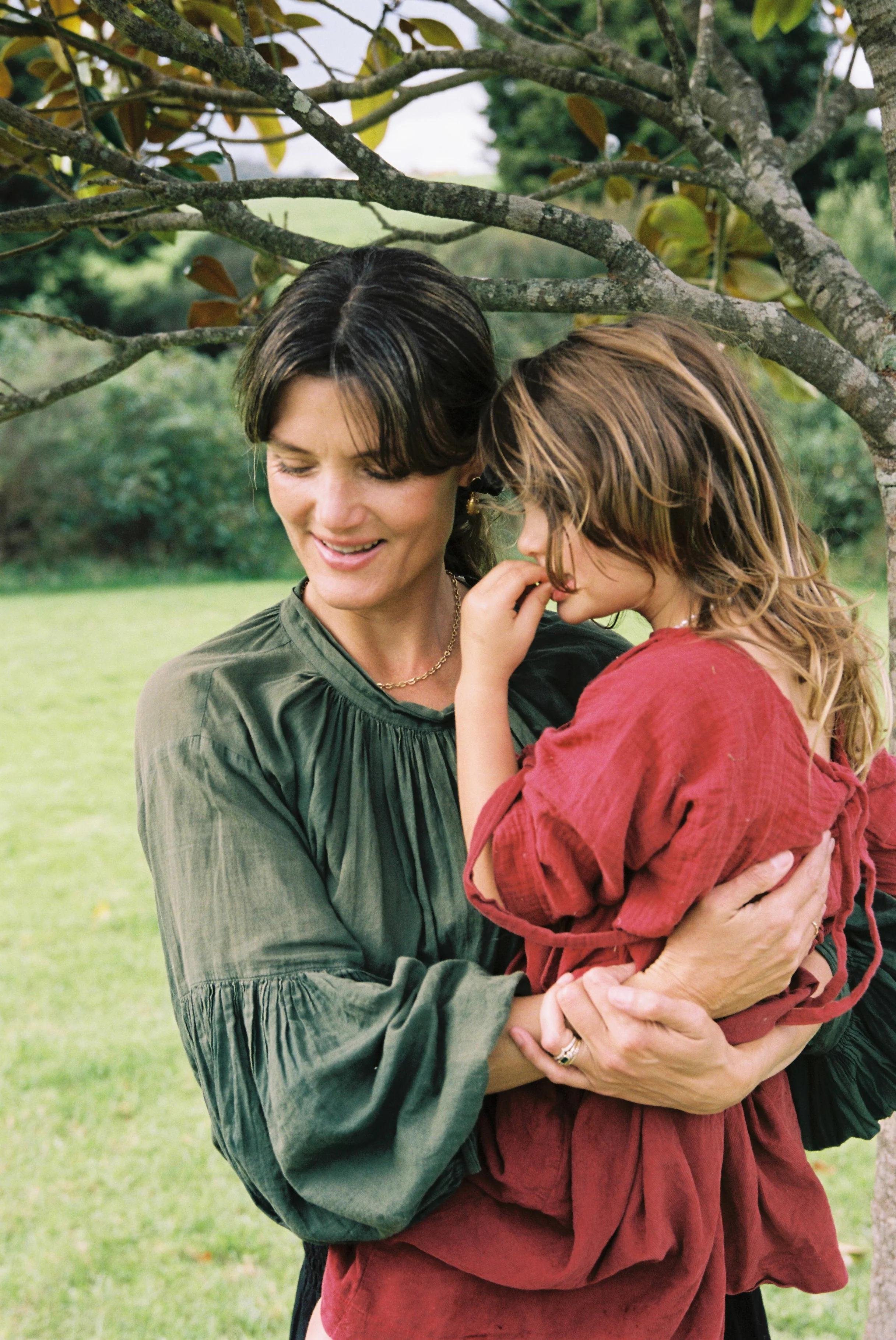 A woman in a green blouse holds a child in a red dress under a tree. She smiles while the child looks away, with one hand near their mouth. Green grass and foliage are in the background.