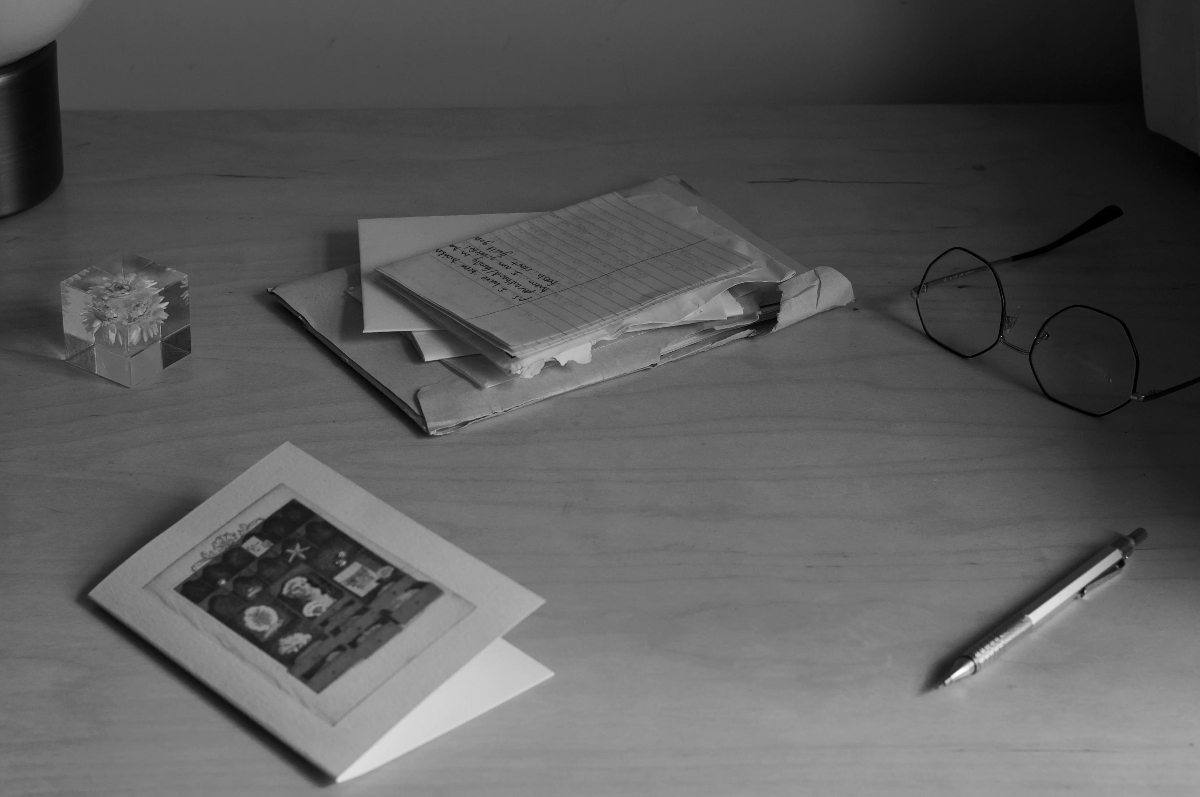 A monochrome image of a desk with an artistic greeting card, a stack of papers with handwritten notes, a pair of glasses, a crystal cube, and a pen.