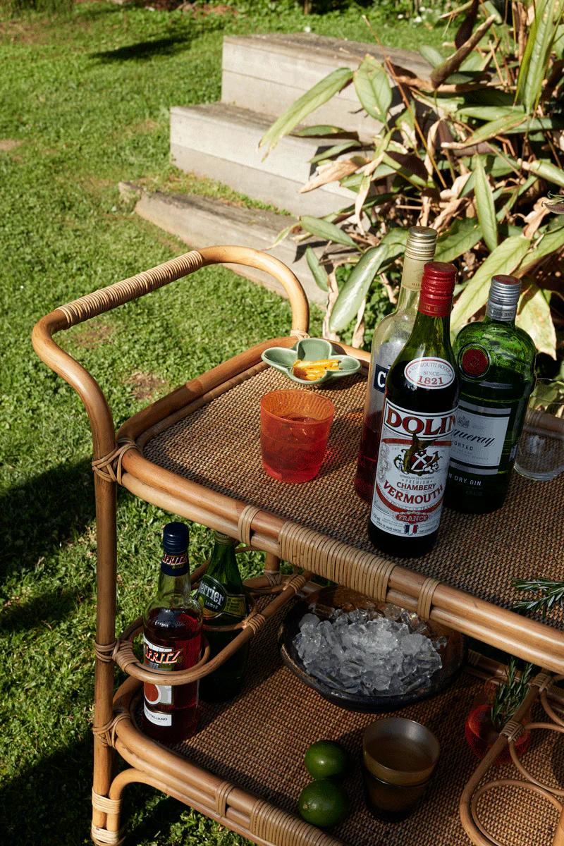 Wicker bar cart on grass with vermouth, gin, and drink ingredients. Steps and plants visible in background.