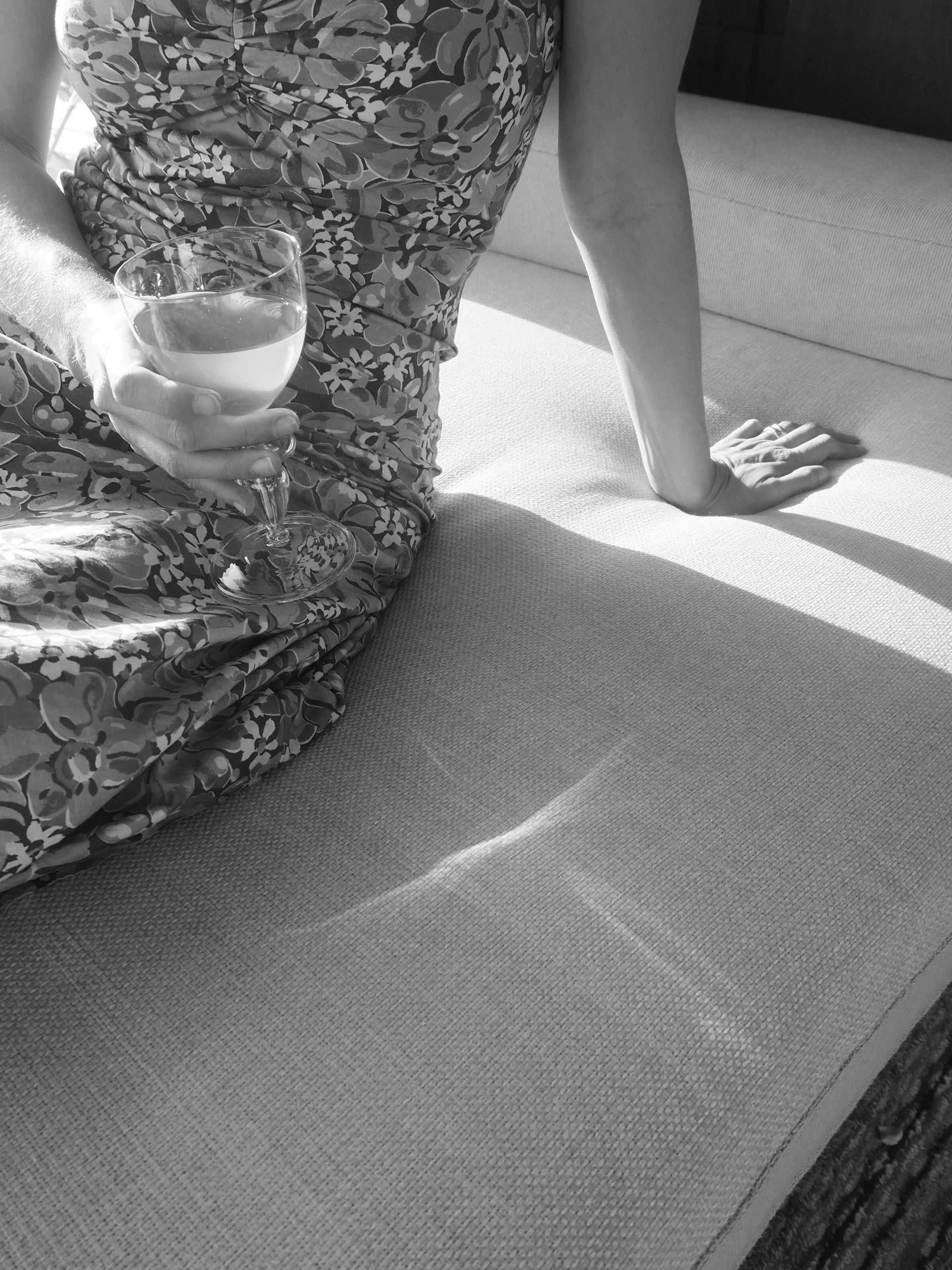 Black and white image: Person in patterned dress holds wine glass while seated on cushion. Sunlight casts shadows. Torso and one arm visible on cushioned seat.