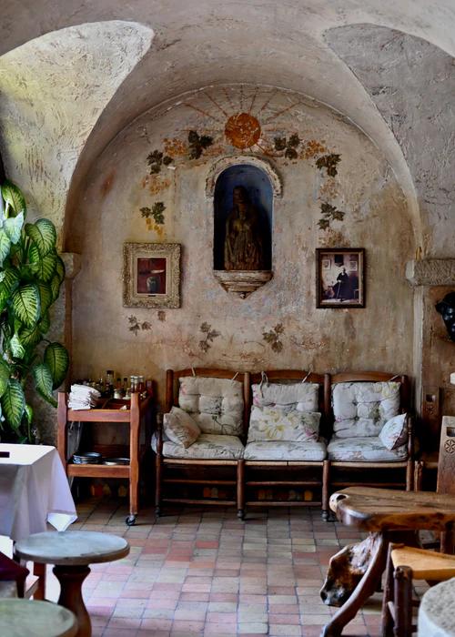 Rustic seating area with arched ceilings and stone alcove housing a small statue. Framed pictures and ivy adorn walls, with wooden chairs nearby.